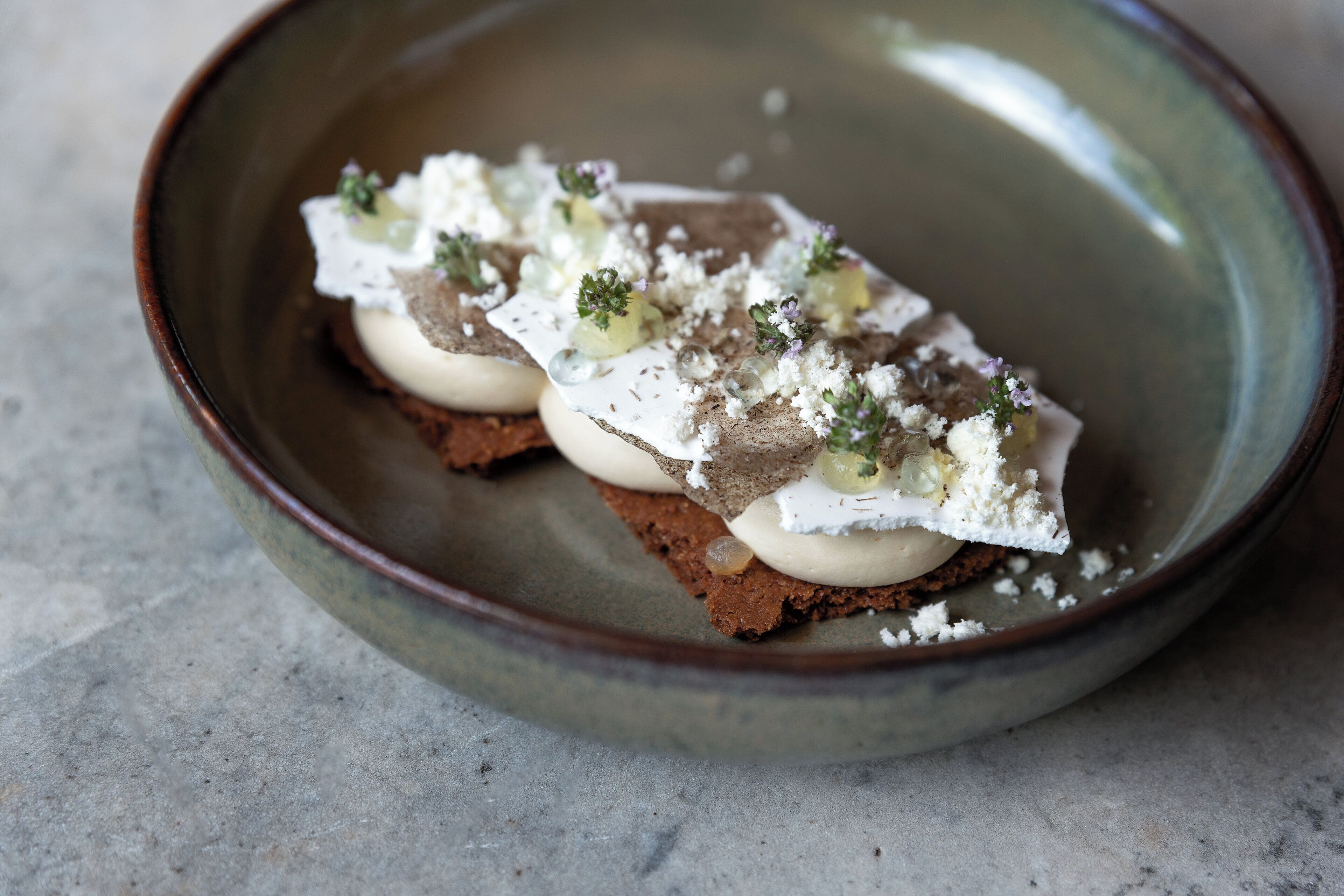 A plate with a dessert consisting of a dark base, white cream, and green toppings.