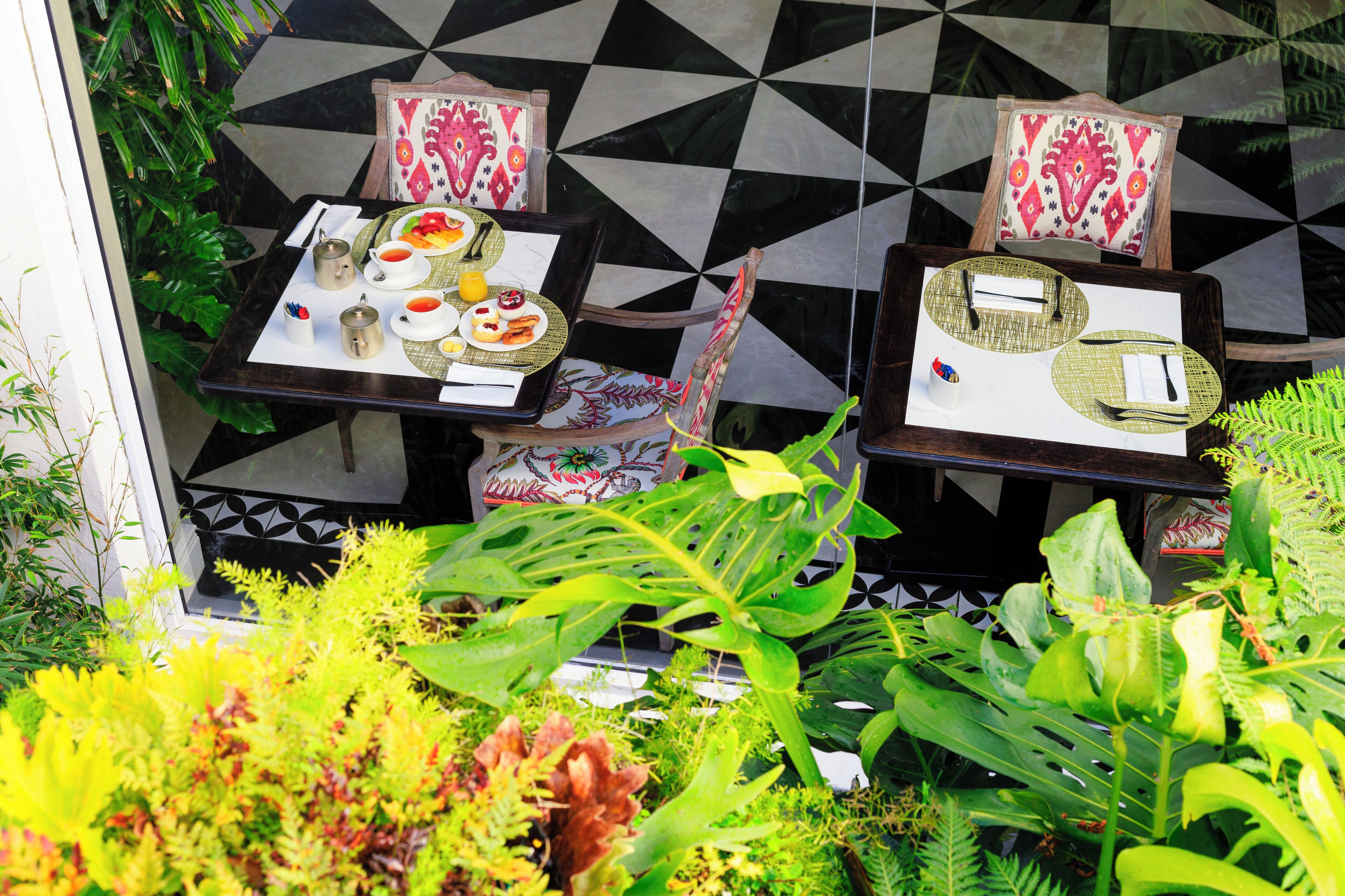 Two tables with placemats, cutlery, and plates with food, set against a geometric tile wall, surrounded by lush greenery.