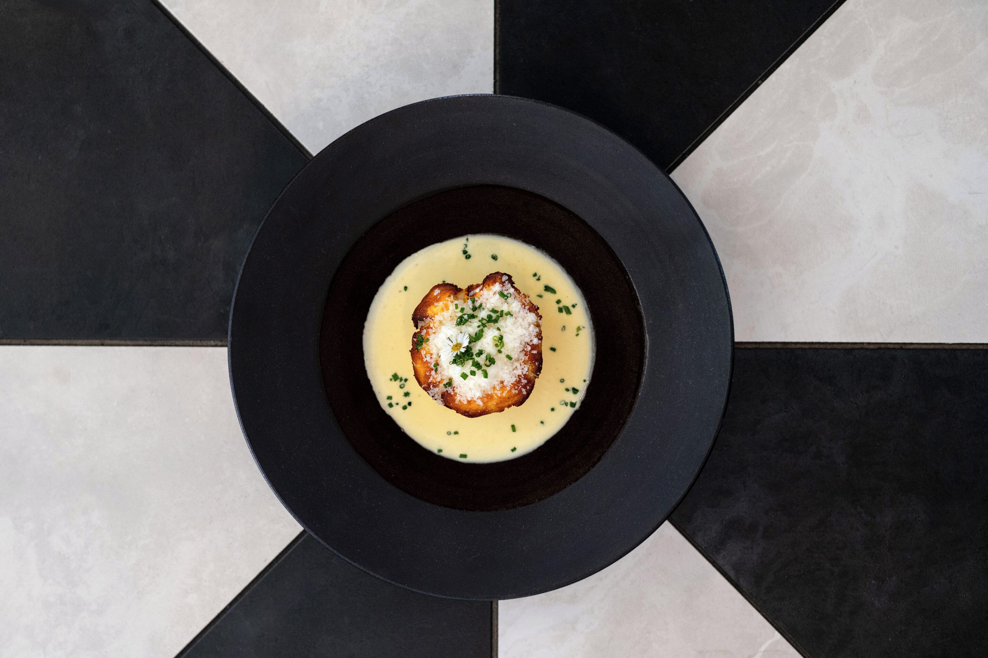 A black plate with a creamy dish and a piece of bread, set on a tiled floor with a geometric pattern.