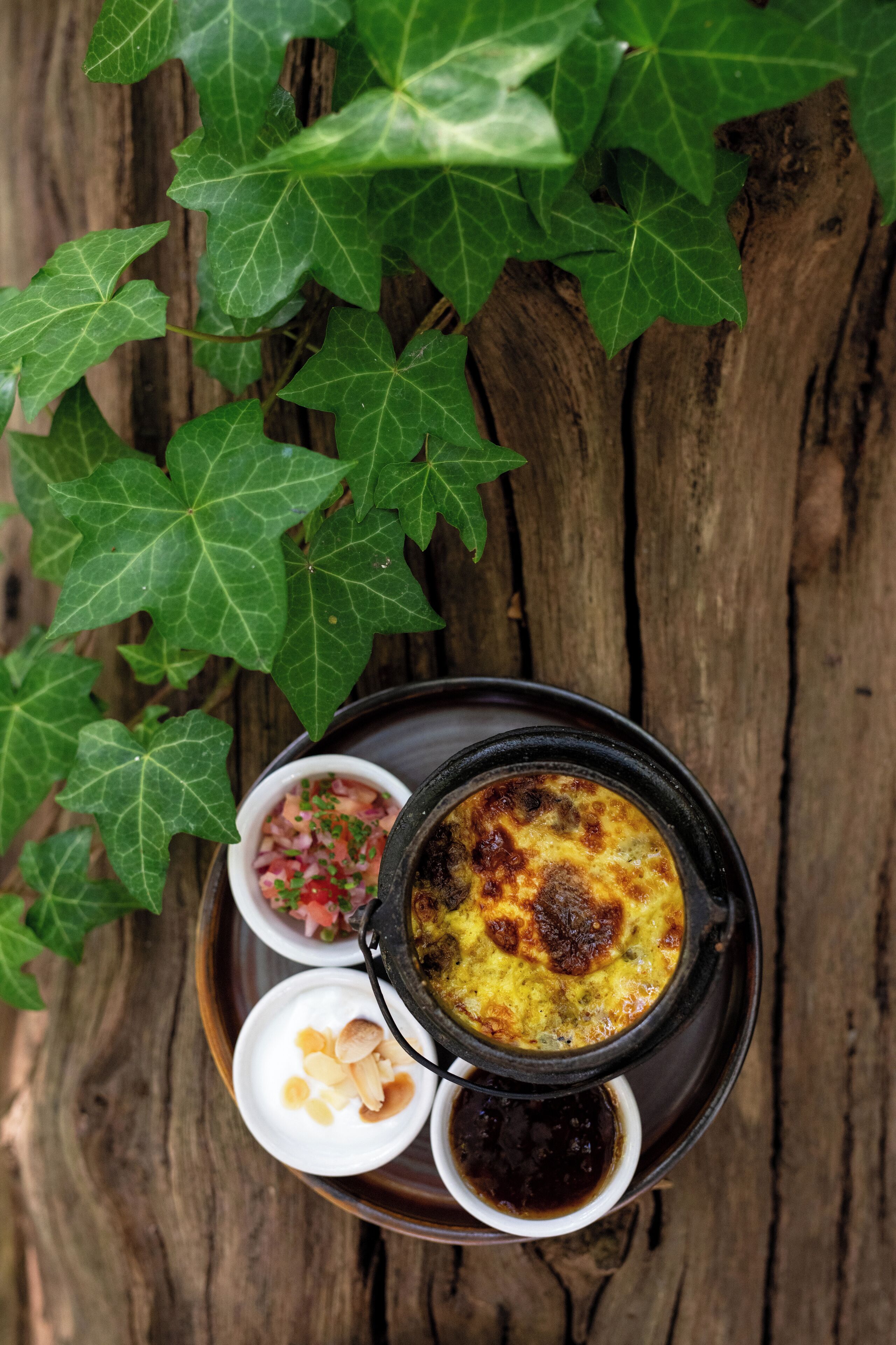 A meal served on a black plate with a bowl of soup, a small bowl of chopped vegetables, and a small bowl of nuts.
