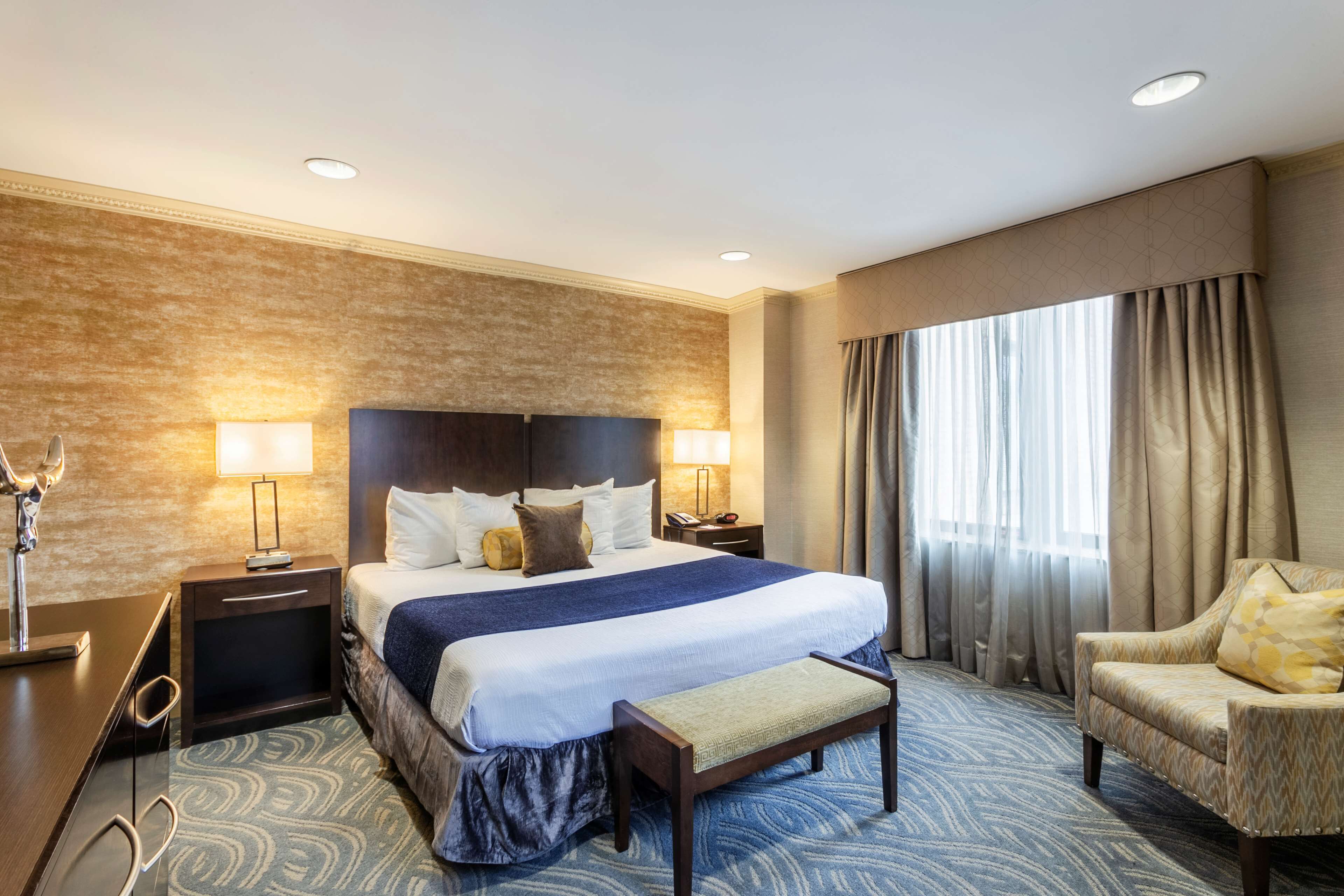 A hotel room with a large bed, two bedside tables with lamps, a chair, and a large window with curtains.