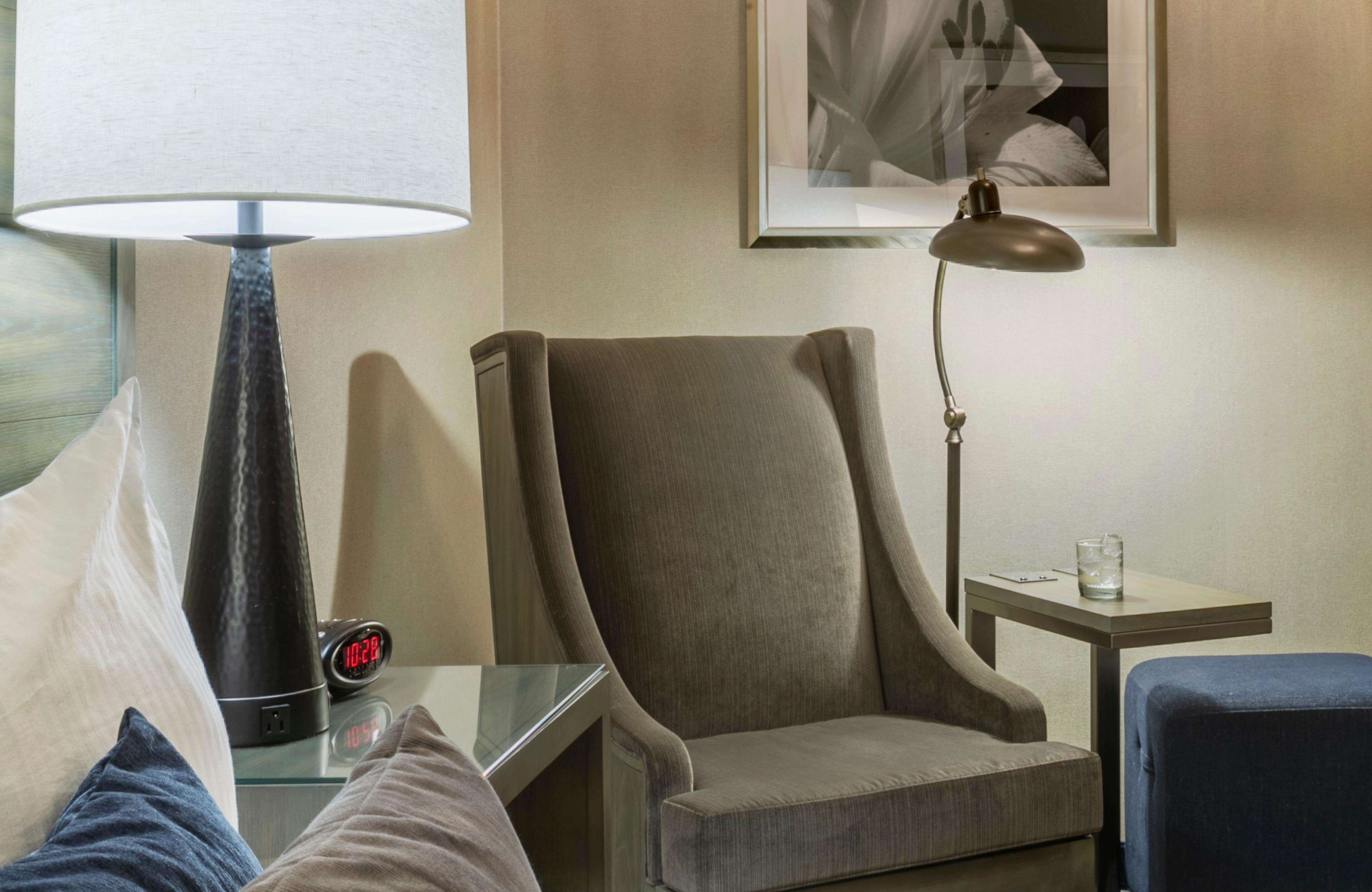 A hotel room with a bed, a chair, a desk, a lamp, and a clock.
