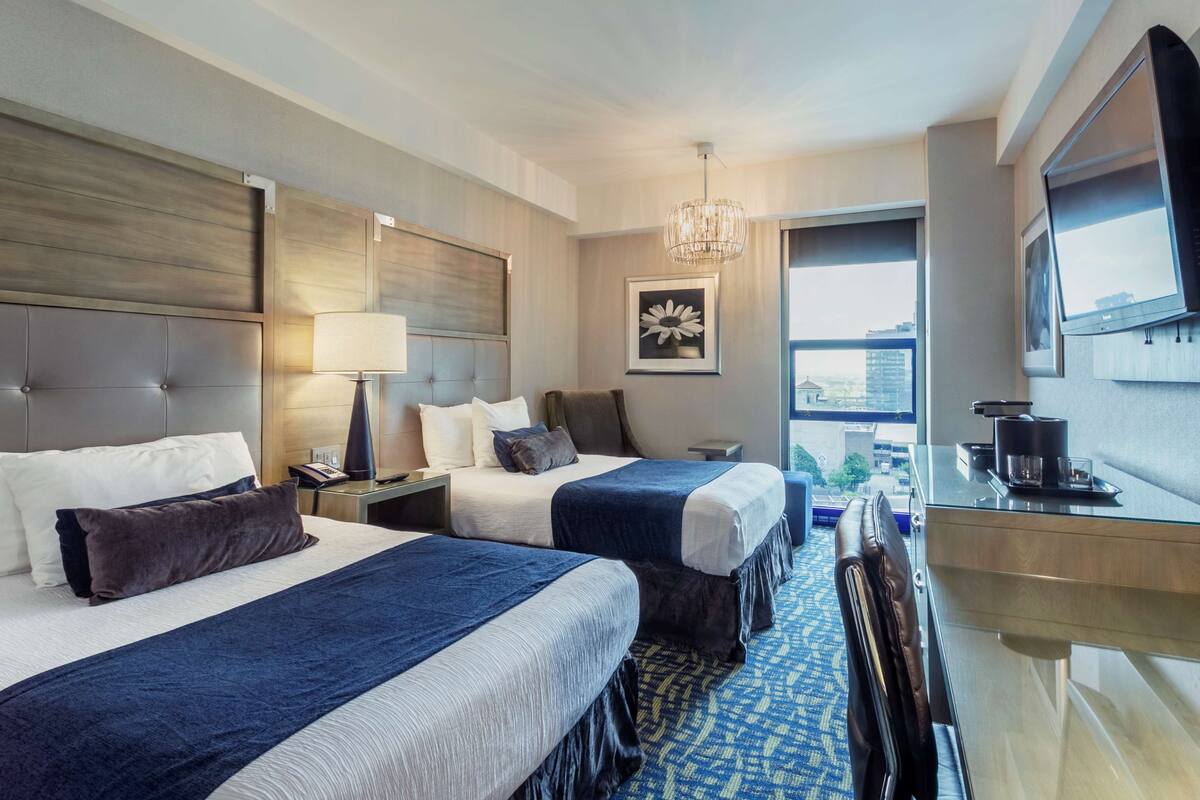 A hotel room with two beds, a desk, a chair, a TV, a window with a city view, and a chandelier.