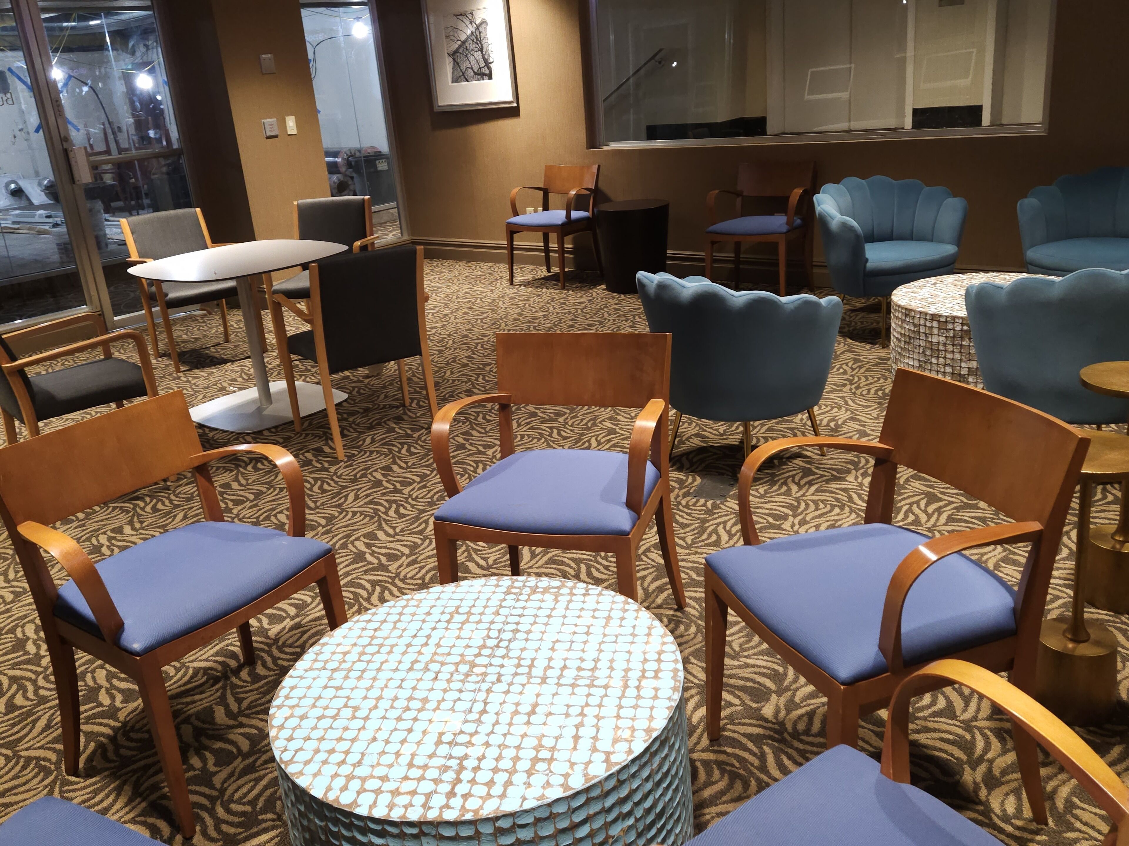 Habitación de hotel con alfombra estampada, sillas de madera con cojines azules, una mesa redonda y un ventanal con vistas al exterior.