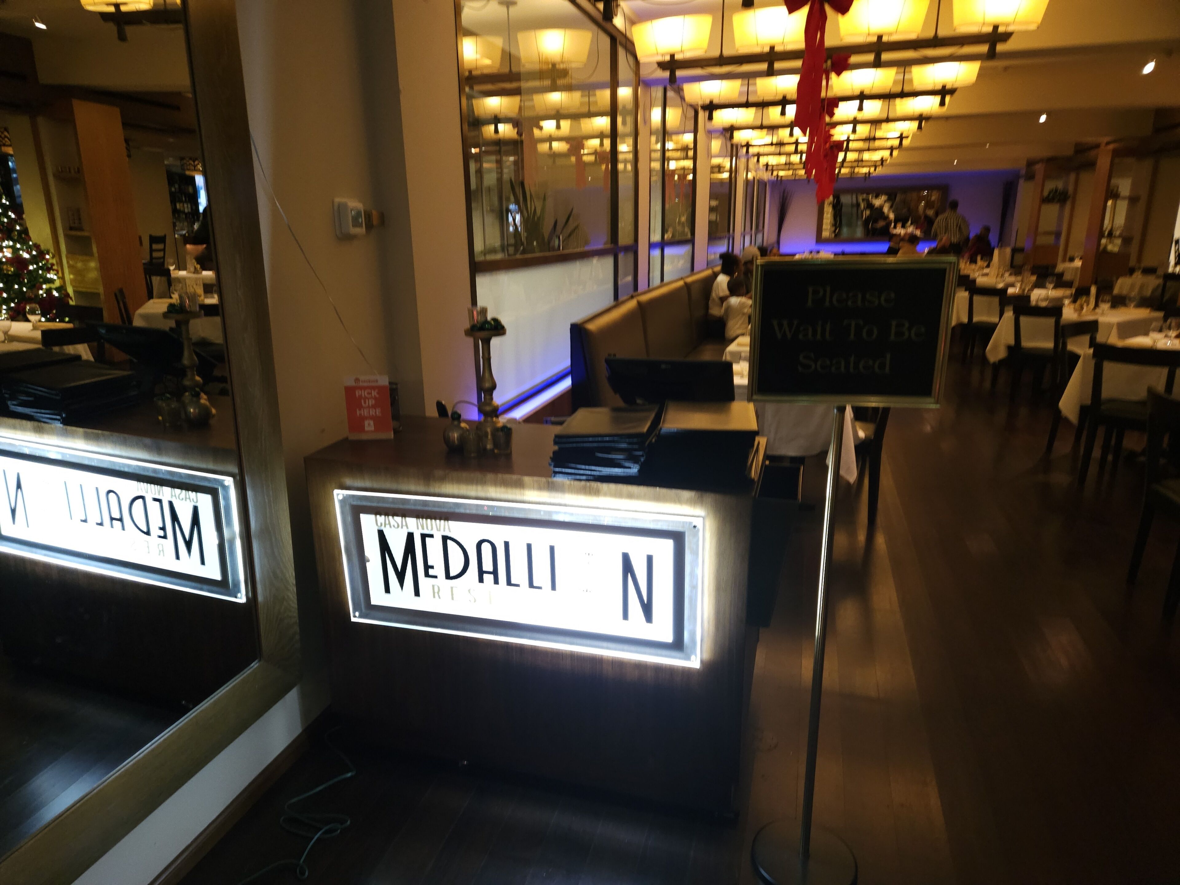 Interior de restaurante com um balcão de recepção identificado como 'MEDALLI N' e uma placa indicando 'RETIRADA AQUI'.