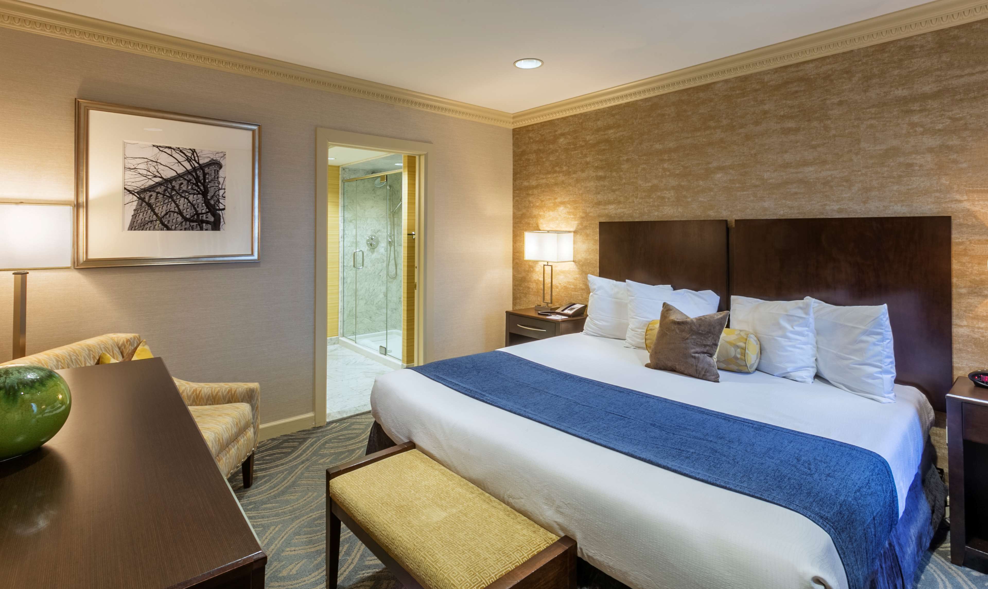 A hotel room with a large bed, a desk, a chair, a framed picture on the wall, and a bathroom visible through an open door.