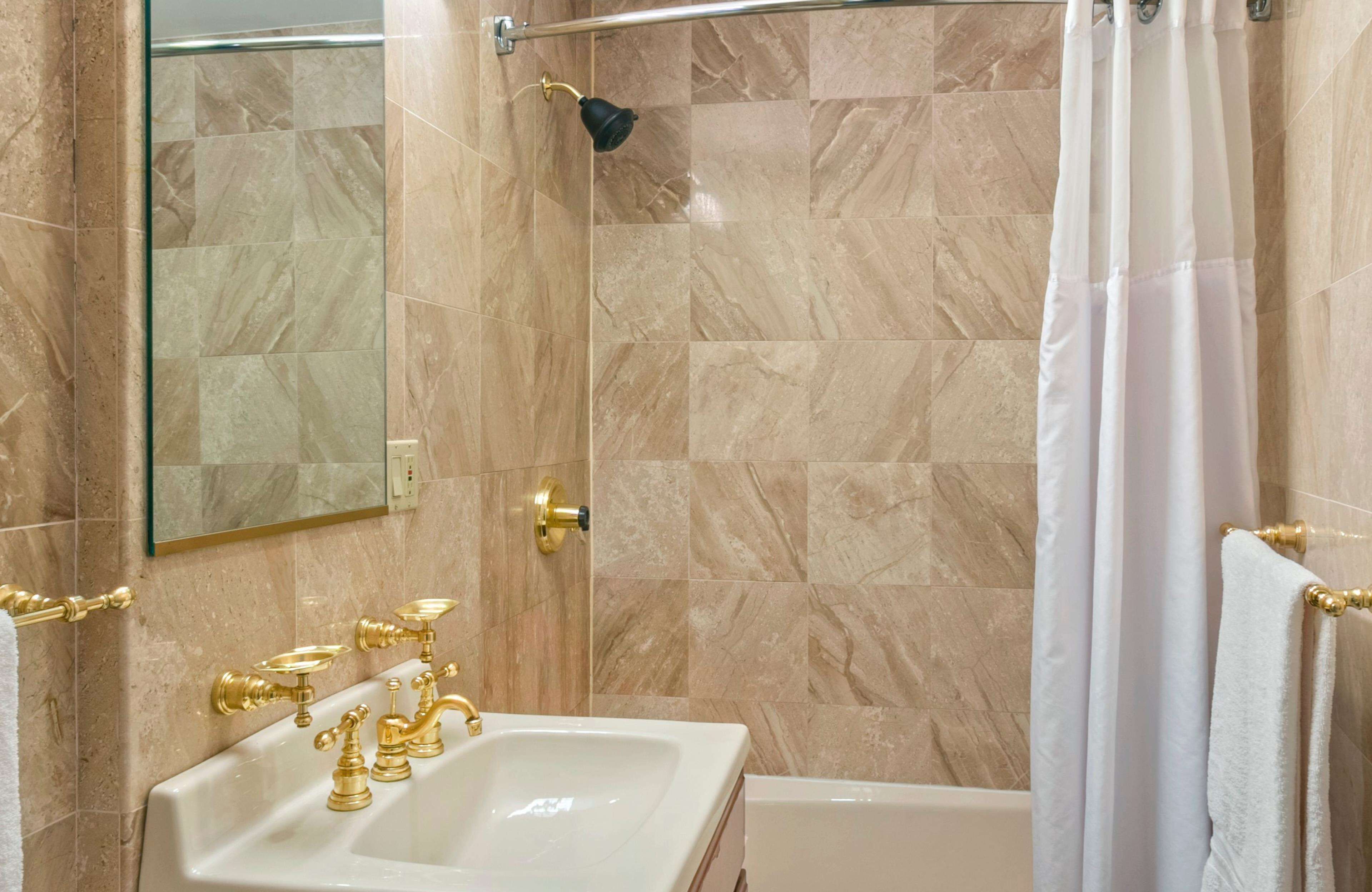 A bathroom with a marble-tiled shower, a white bathtub, and a sink with gold fixtures.