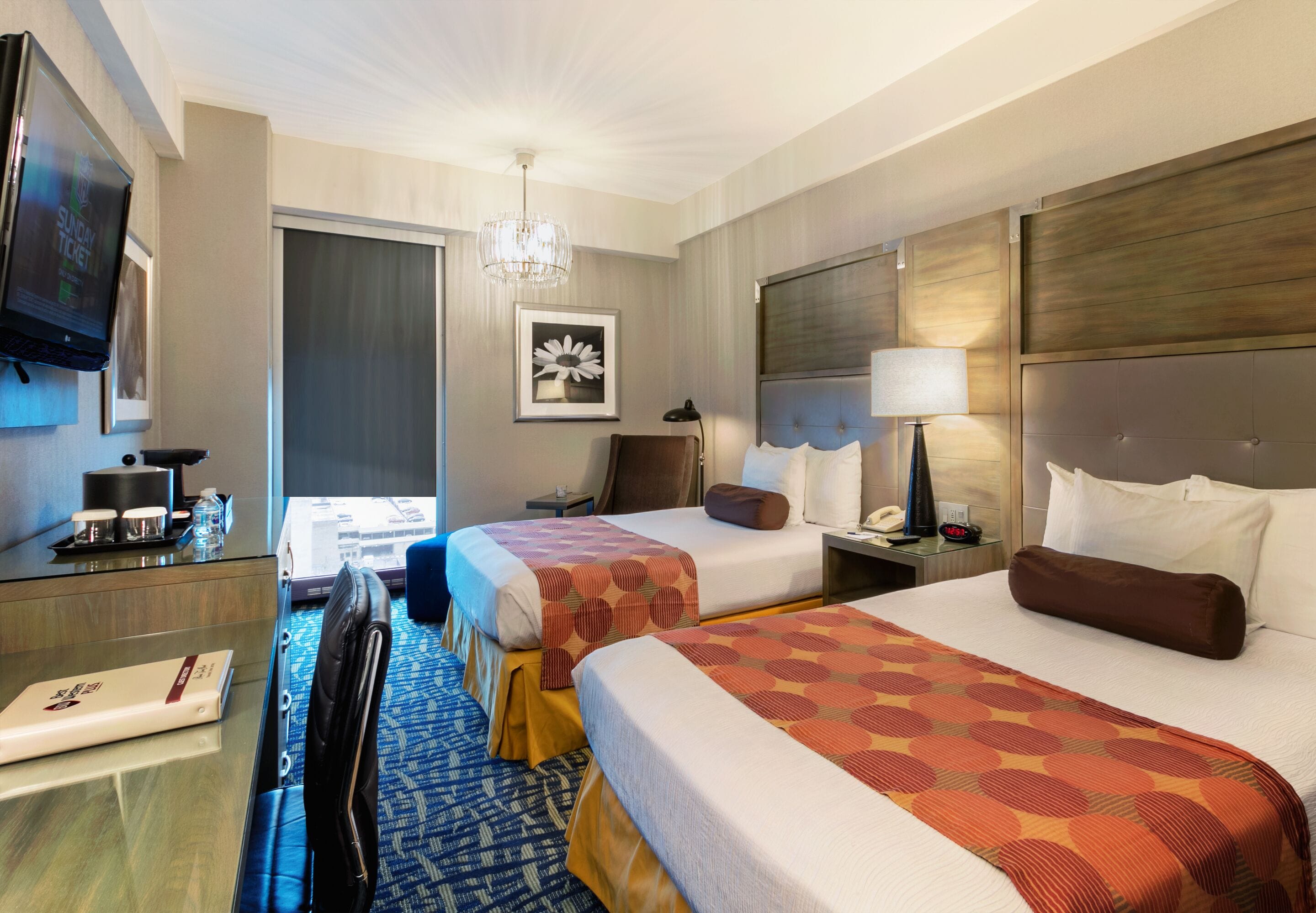 Quarto de hotel com duas camas, uma escrivaninha, uma cadeira, uma televisão, um candelabro e uma janela com cortinas.