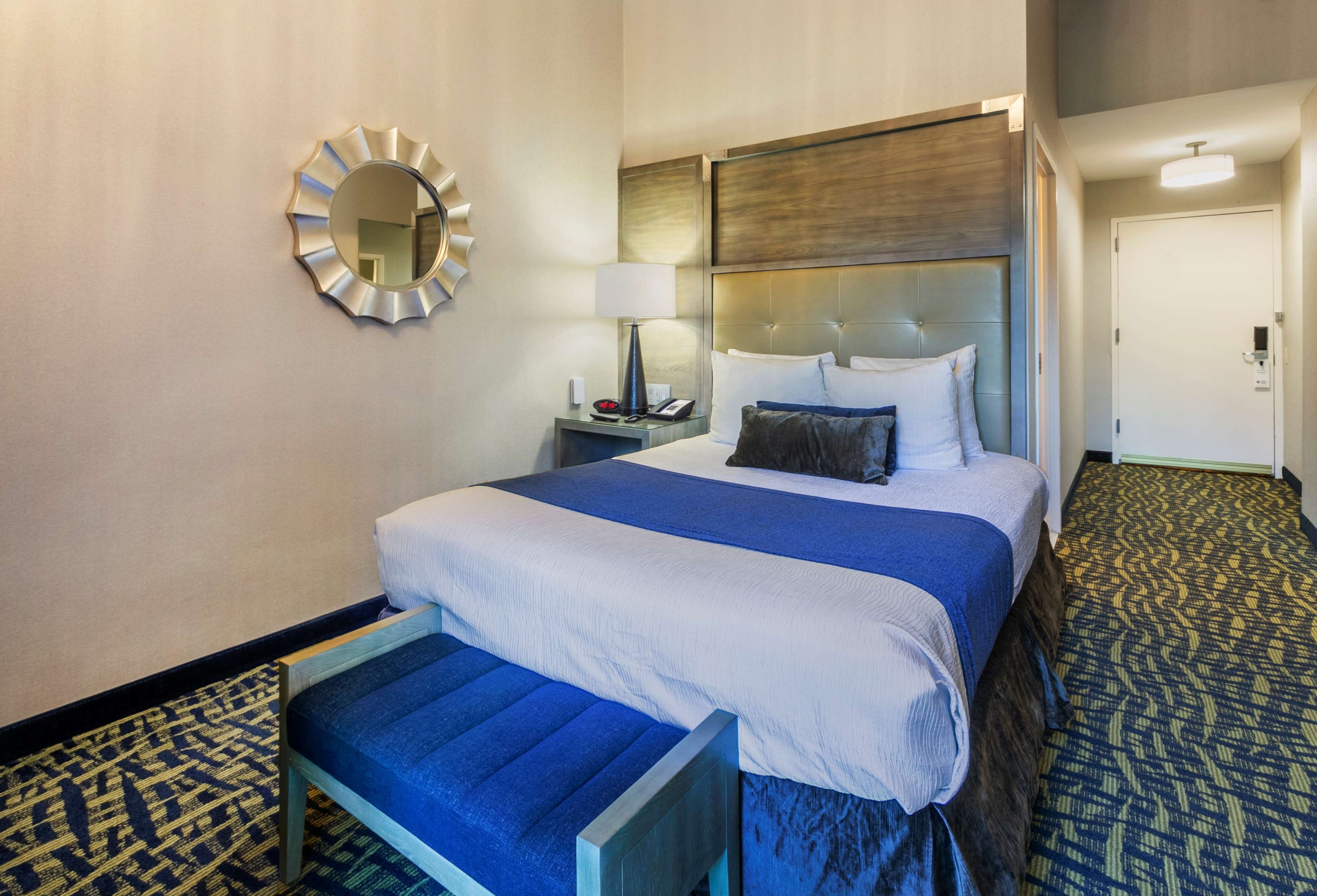 Une chambre d’hôtel avec un grand lit, une table de chevet avec une lampe, un miroir au mur et une moquette à motifs.