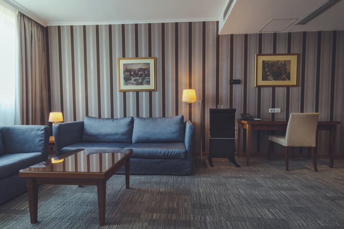 A hotel room with a striped wallpaper, a blue sofa, a wooden coffee table, a desk with a lamp, and two framed pictures on the wall.