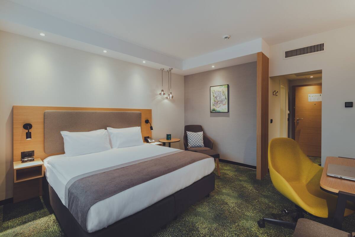 A hotel room with a large bed, bedside tables, a desk, a chair, and a painting on the wall.