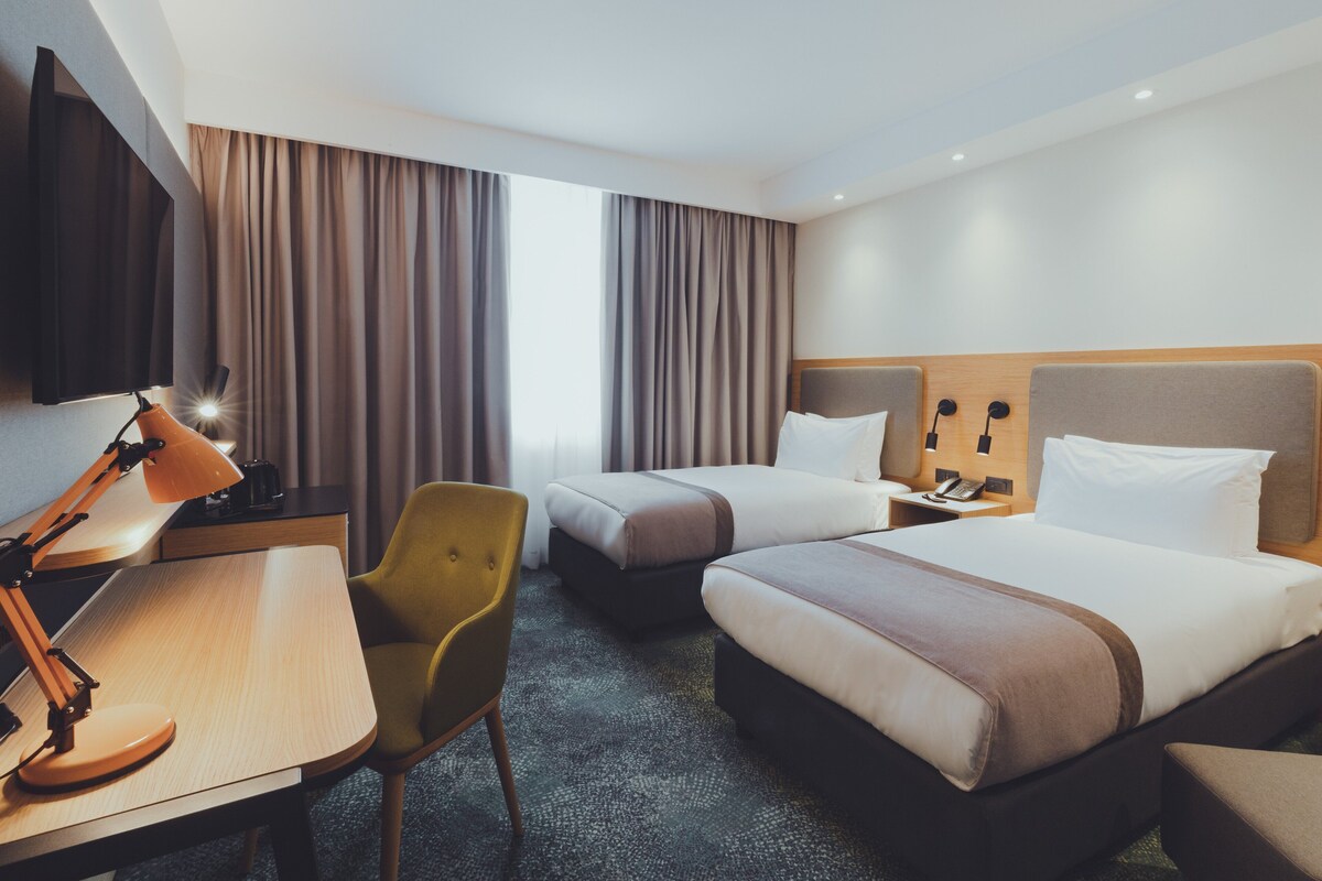 A hotel room with two beds, a desk with a lamp, a chair, and a large window with curtains.