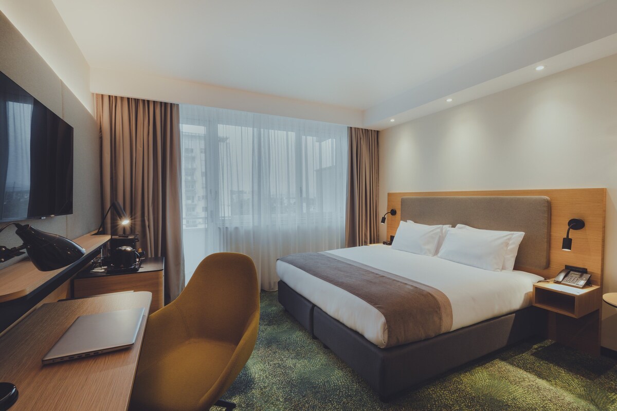 A hotel room with a large bed, a desk with a laptop, a chair, a TV, and a bedside table with a phone.
