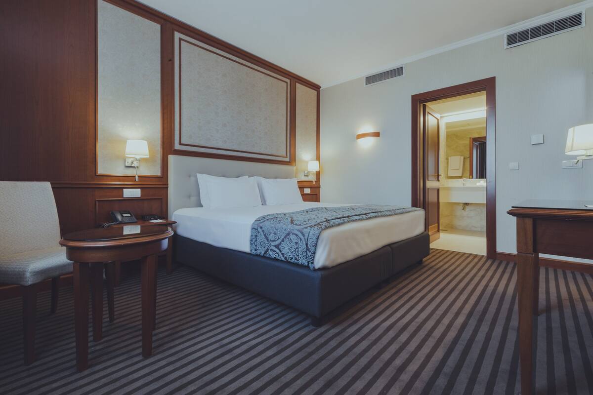 A hotel room with a large bed, a bedside table, a chair, and a desk.