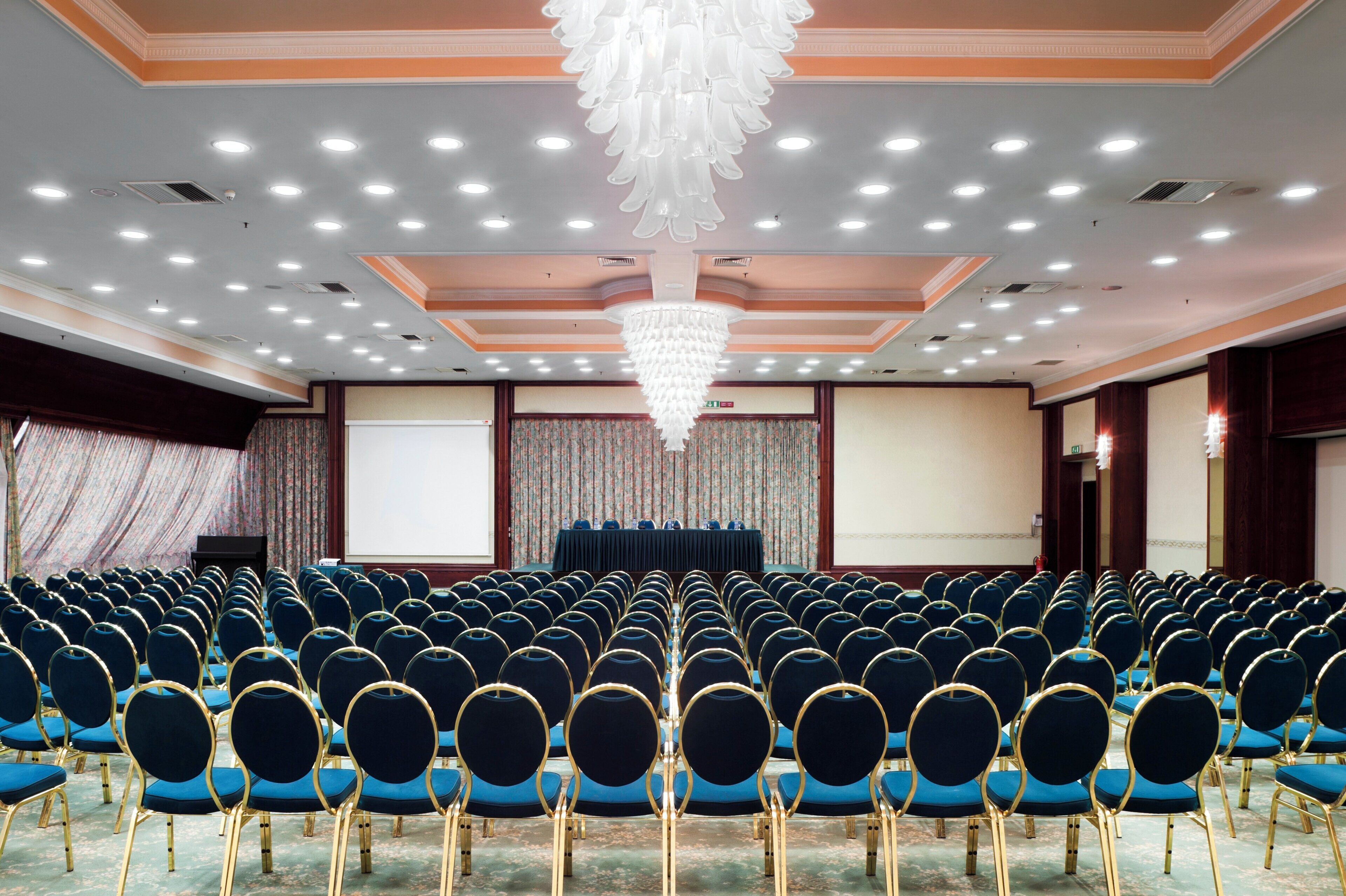 Een congreszaal met rijen blauwe stoelen, een podium met een scherm en een kroonluchter.