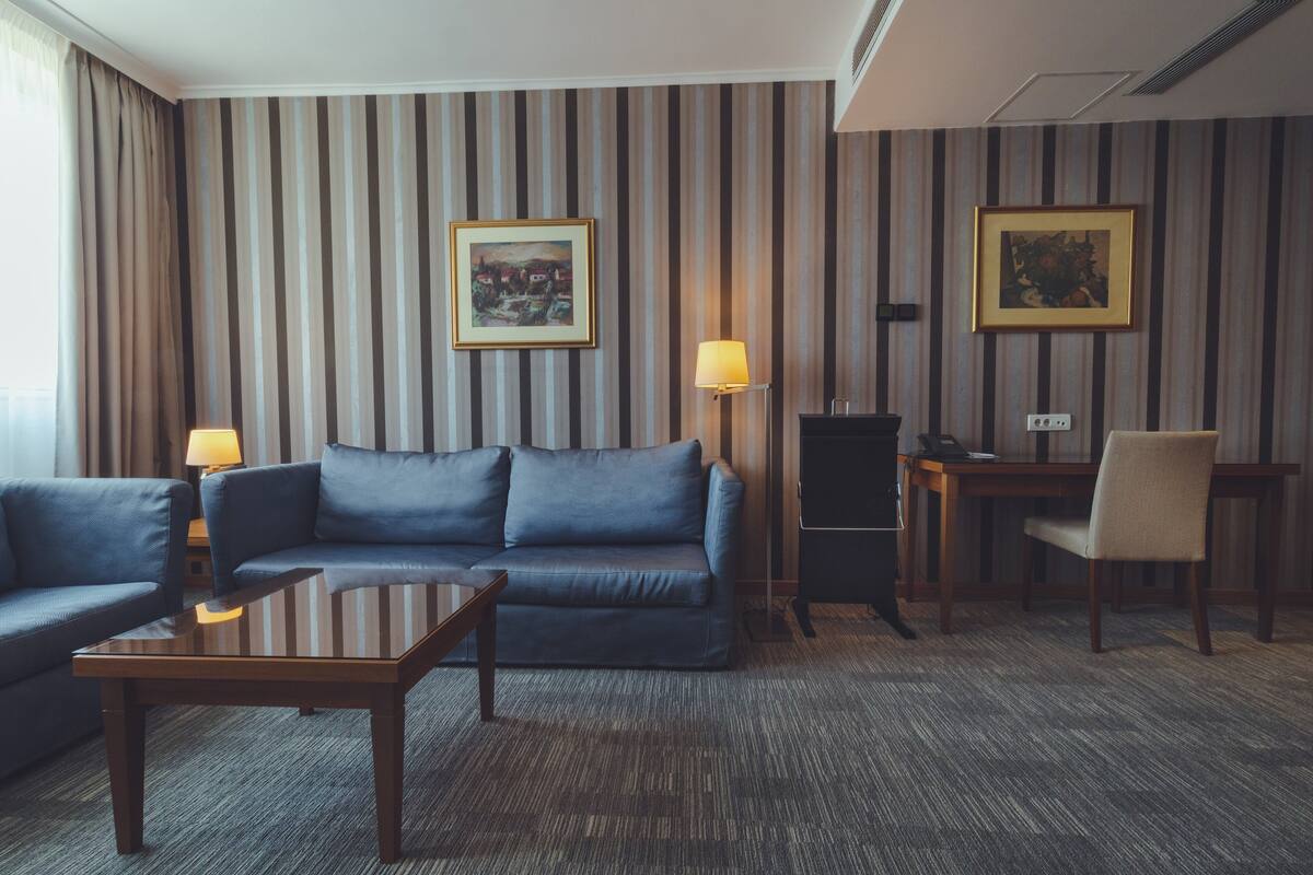 A hotel room with a striped wallpaper, a blue sofa, a wooden coffee table, a desk with a lamp, and two framed pictures on the wall.