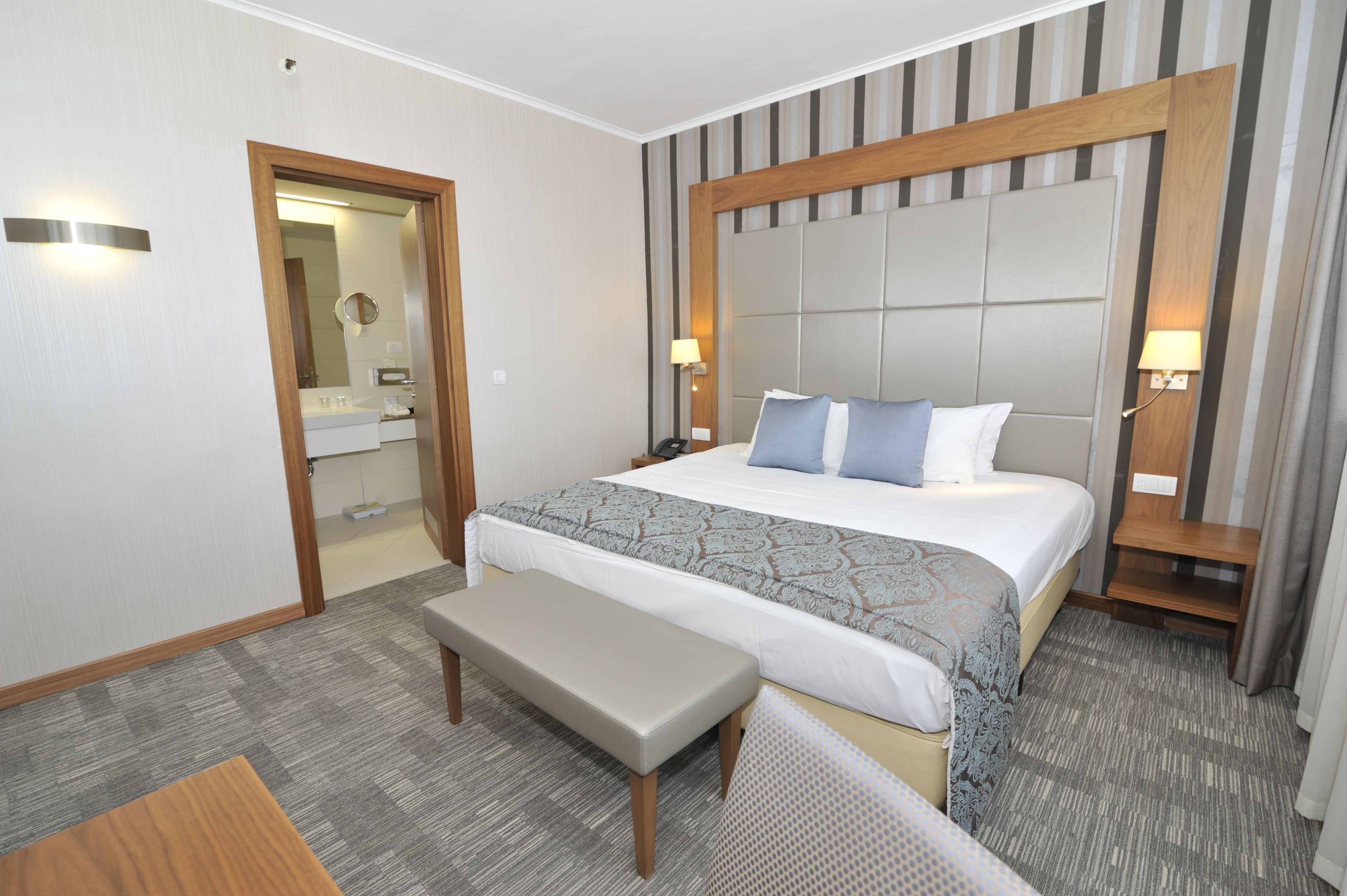 A hotel room with a large bed, bedside tables, a bench, a mirror, and a door.
