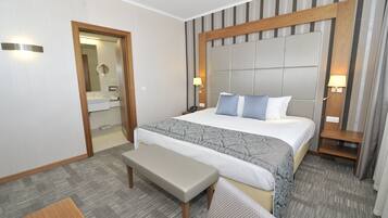 A hotel room with a large bed, bedside tables, a bench, a mirror, and a door.