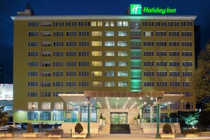 A multi-story Holiday Inn hotel with a prominent green logo, a covered entrance, and a parking area with cars. - Holiday Inn Skopje by IHG (Skopje)