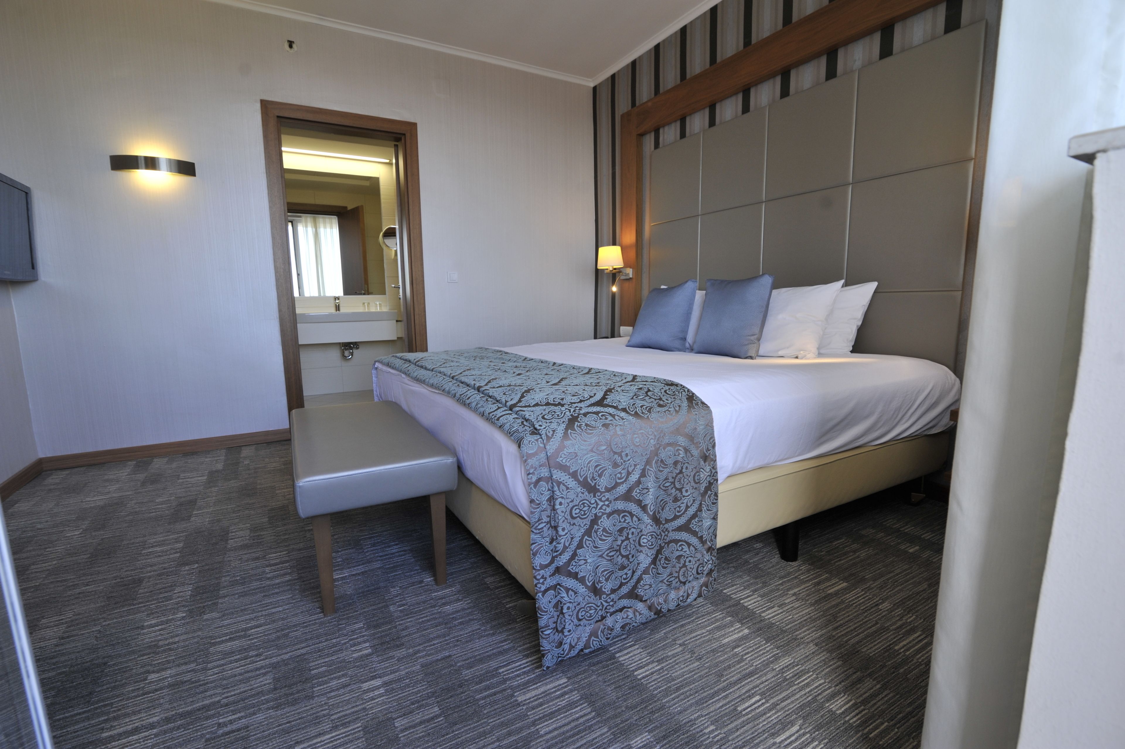 Habitación de hotel con una cama grande, una mesita de noche, una silla, un espejo y un televisor instalado en la pared.
