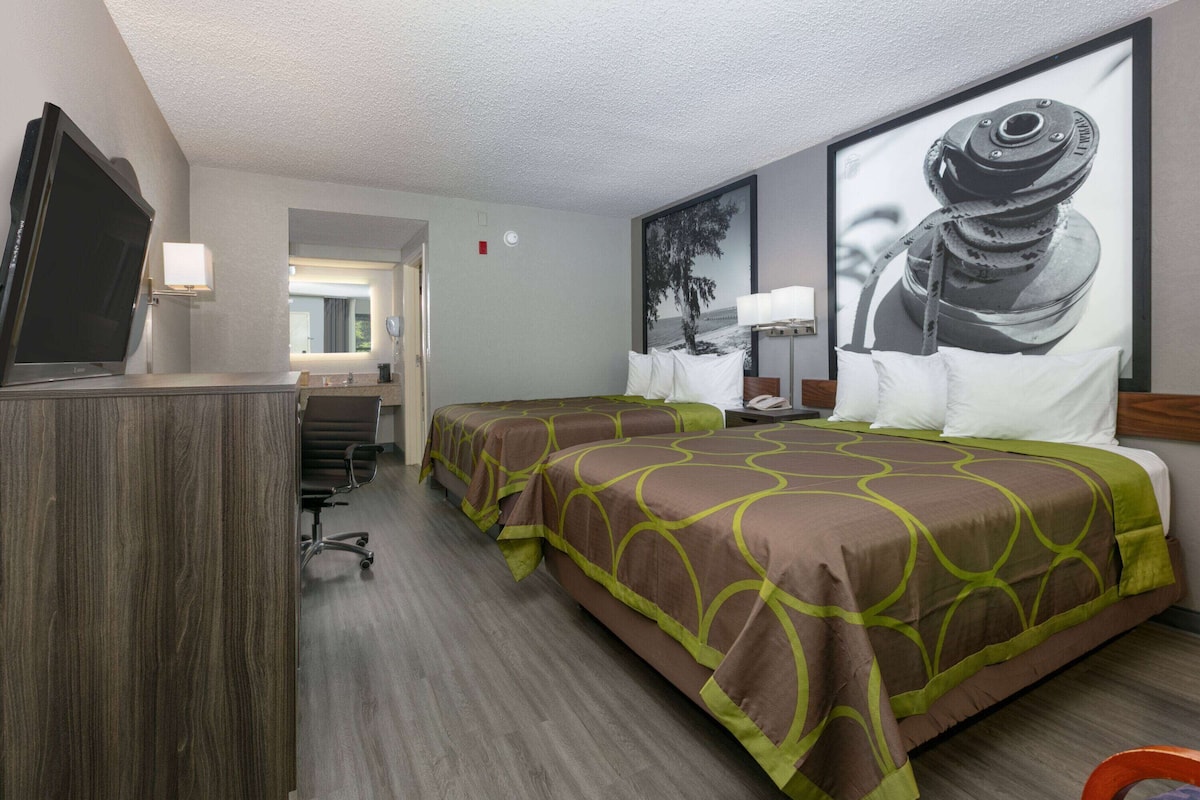 Quarto de hotel com duas camas, uma escrivaninha com computador, televisão e um quadro grande na parede.