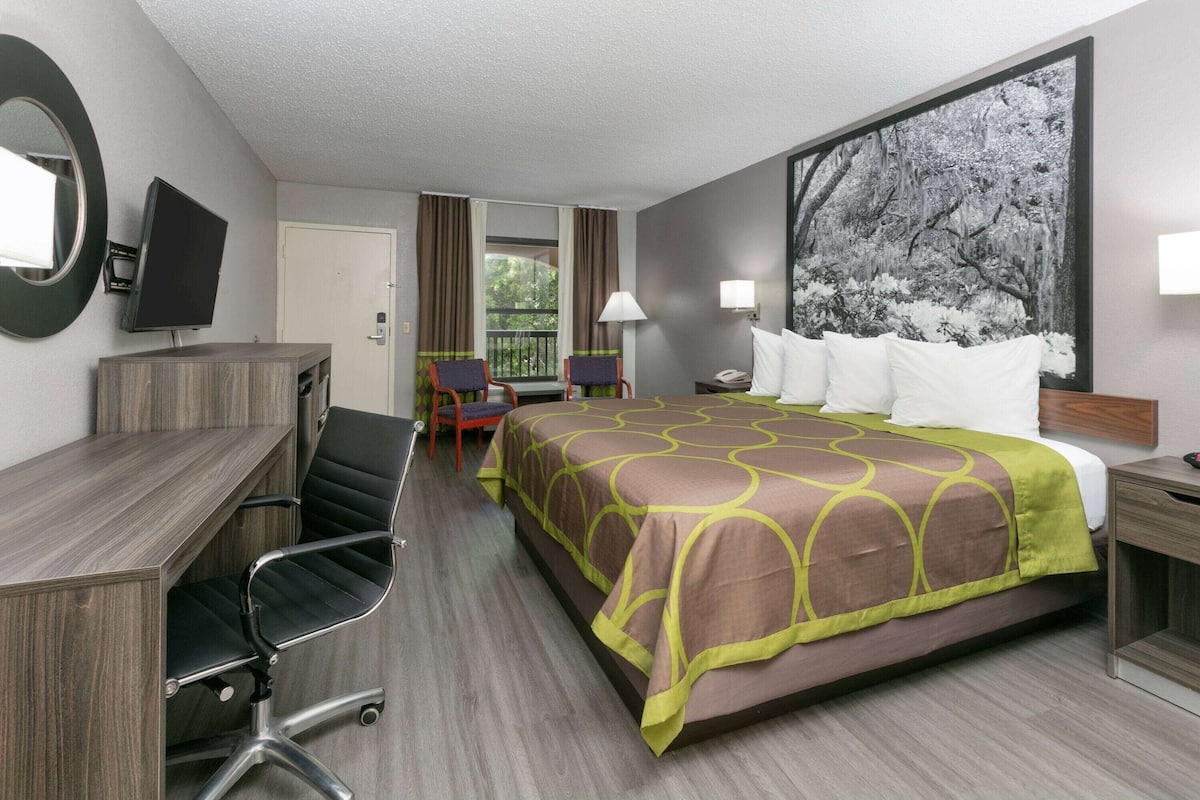 Quarto de hotel com cama, escrivaninha, cadeira, televisão e varanda com cortinas.