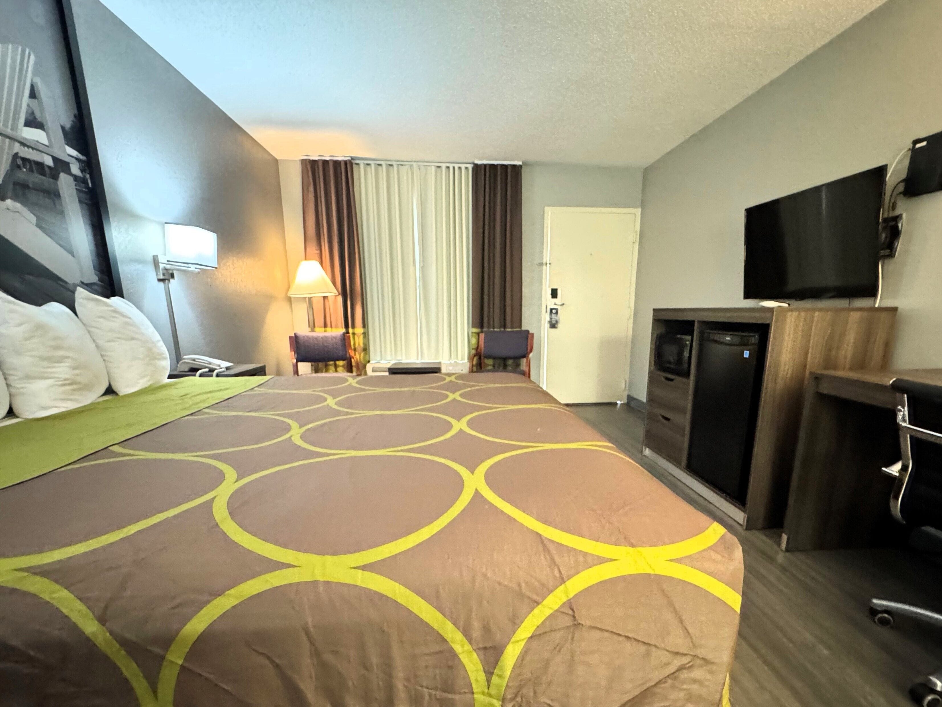 A hotel room with a large bed, a desk with a television, a chair, and a lamp.