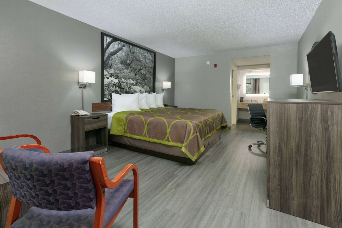 Quarto de hotel com cama, escrivaninha, cadeira, televisão e um quadro na parede.