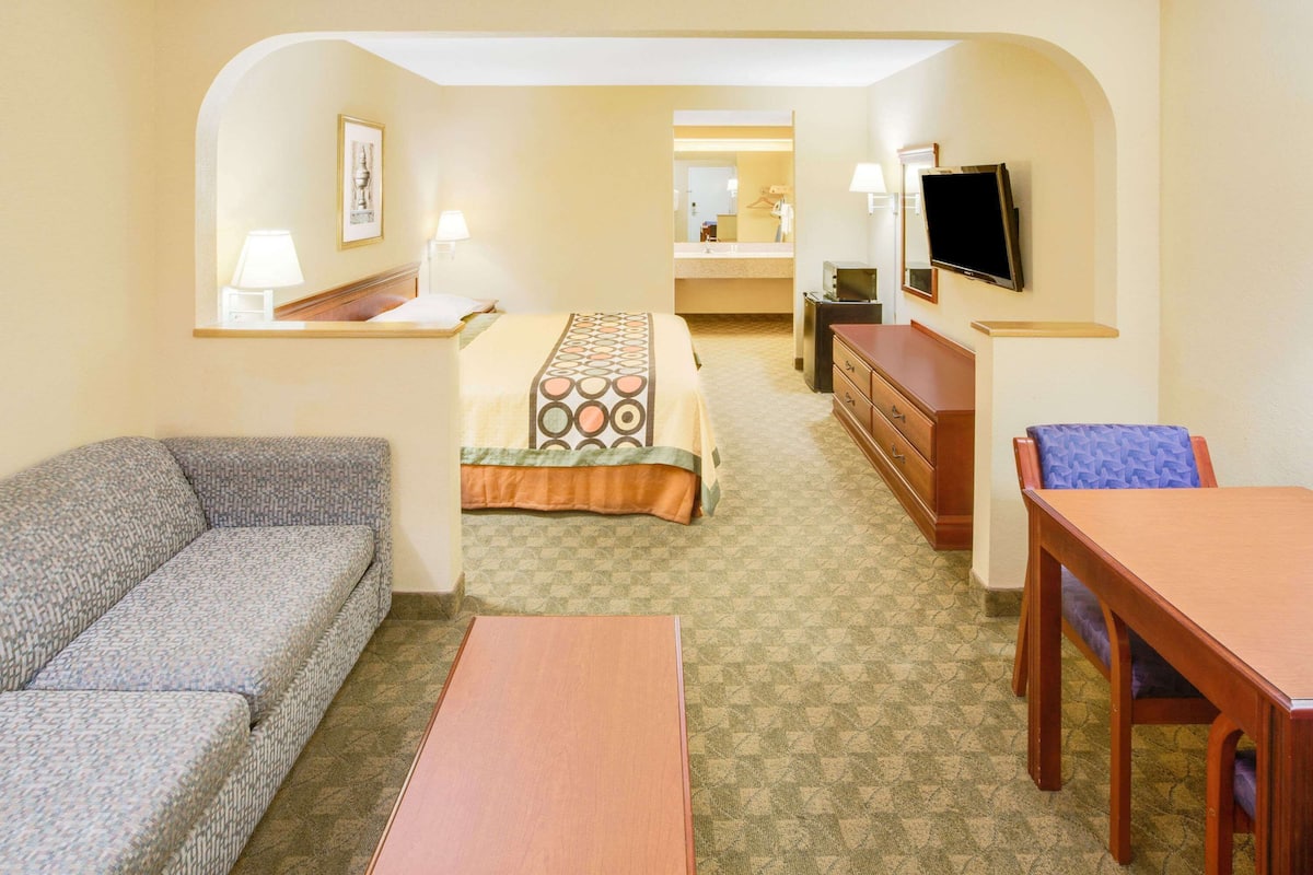 Quarto de hotel com sofá, cama com colcha estampada, penteadeira de madeira, televisão e banheiro visível por uma porta arqueada.