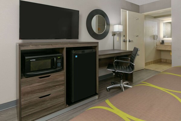 Habitación de hotel con cama, escritorio con microondas, refrigerador, silla, espejo y puerta que da a un baño.