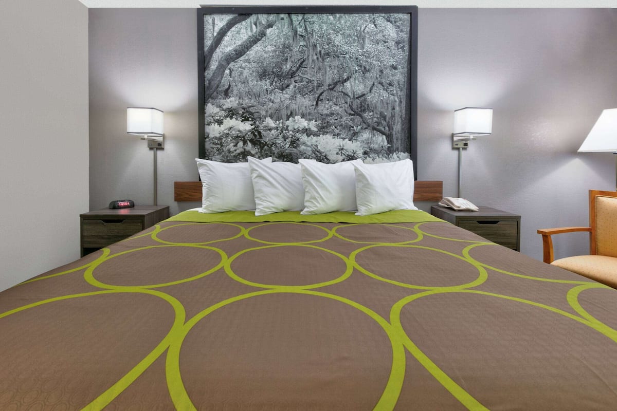 Quarto de hotel com uma cama grande, duas mesas de cabeceira, um criado-mudo, uma cadeira e uma luminária fixada na parede.