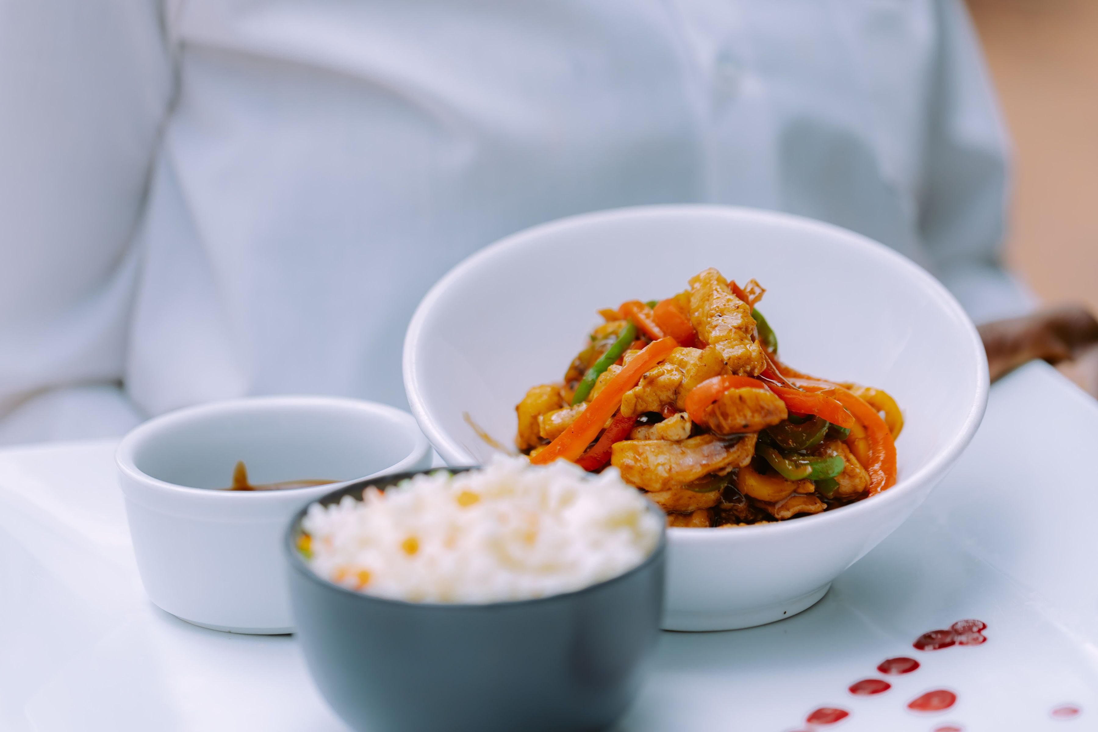 A bowl of stir-fried vegetables and shrimp, a small bowl of rice, and a small bowl of sauce on a white table.