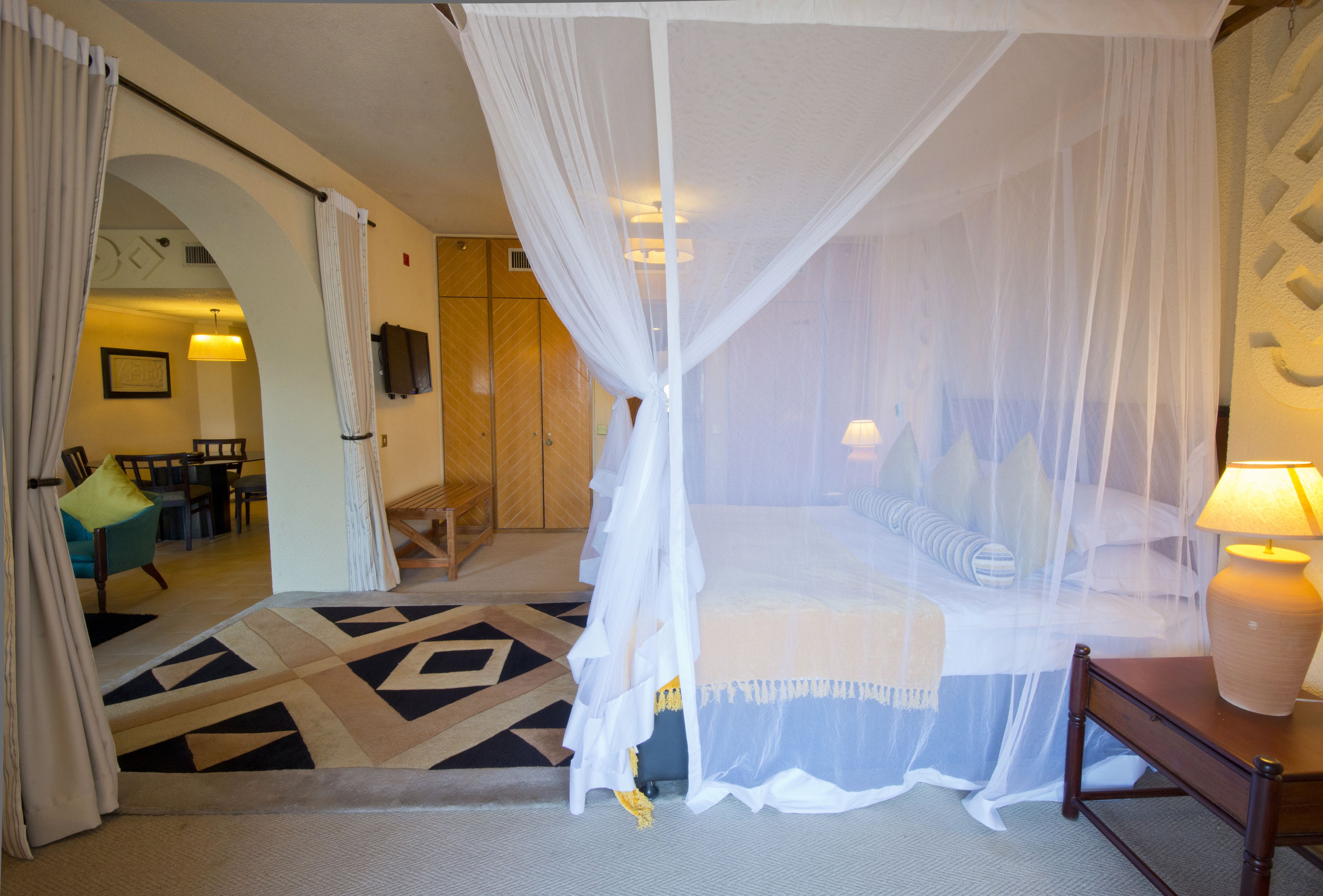 Una habitación de hotel con una cama bajo un dosel, una mesita de noche con una lámpara, una alfombra estampada y vistas al interior del hotel.