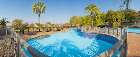 A swimming pool with a blue tiled border, surrounded by lounge chairs and thatched umbrellas, with palm trees and a clear sky in the background.