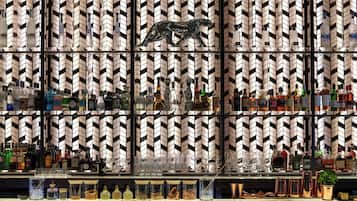 Una zona de bar con estantes llenos de botellas de licor y vasos, una escultura metálica decorativa y una pared con diseño.
