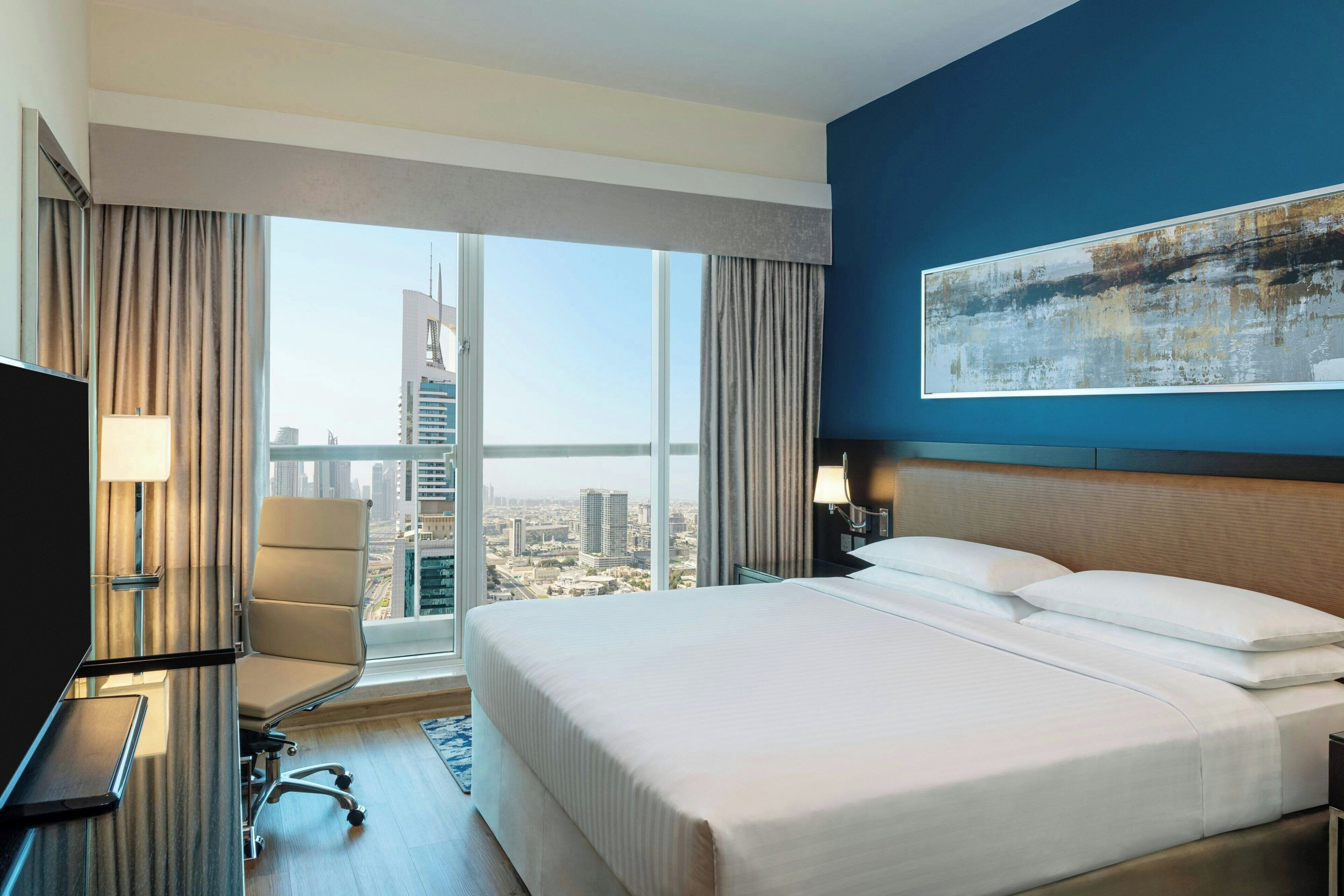 Habitación de hotel con una cama grande, un escritorio con silla, televisión y vistas a la ciudad a través de amplios ventanales.