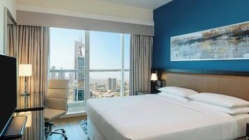 Habitación de hotel con una cama grande, un escritorio con silla, televisión y vistas a la ciudad a través de amplios ventanales.