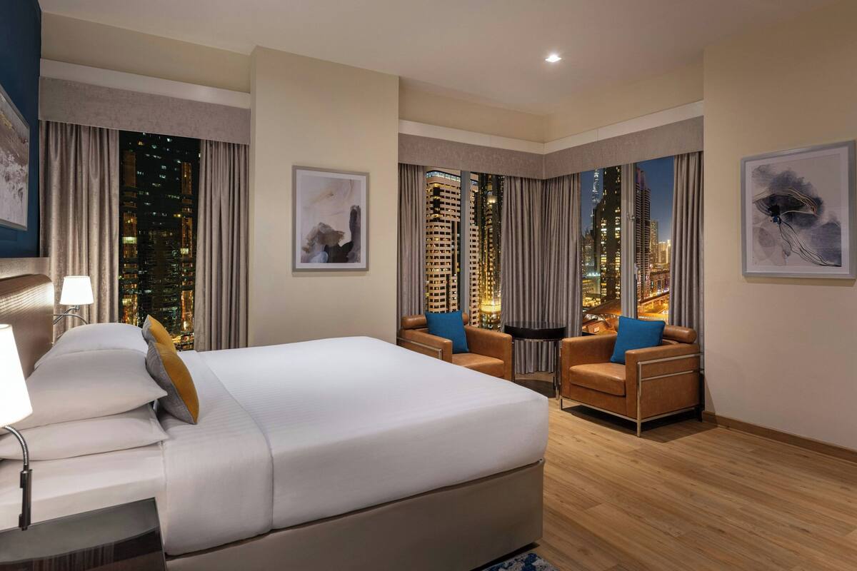 A hotel room with a large bed, two armchairs, a nightstand, and a view of a cityscape at night.