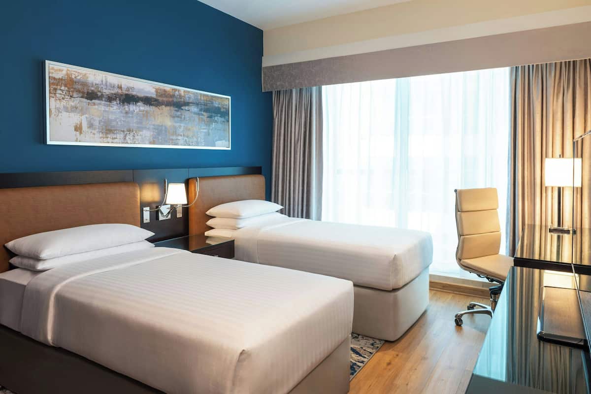 A hotel room with two beds, a desk, a chair, a lamp, and a large window with curtains.