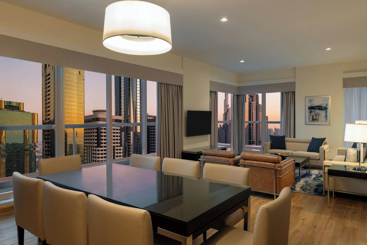 A modern hotel room with a large dining table, comfortable seating, a flat-screen TV, and large windows offering city views.