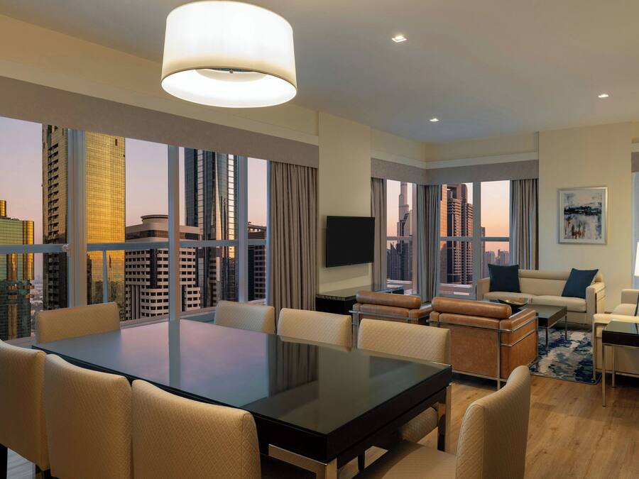 A modern hotel room with a large dining table, comfortable seating, a flat-screen TV, and large windows offering city views.