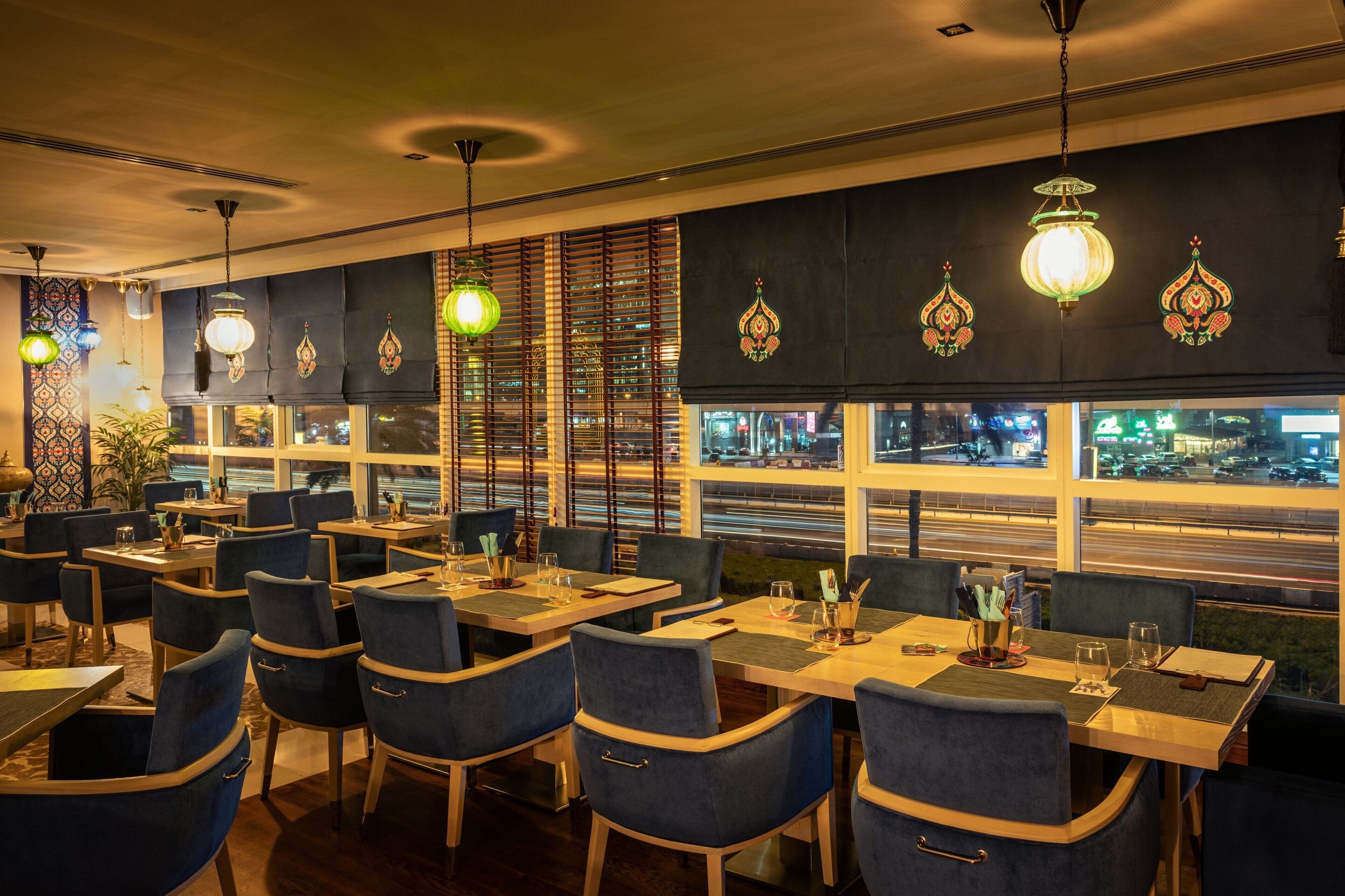 Un restaurante con mesas y sillas, ventanas amplias con persianas y luces decorativas colgantes.