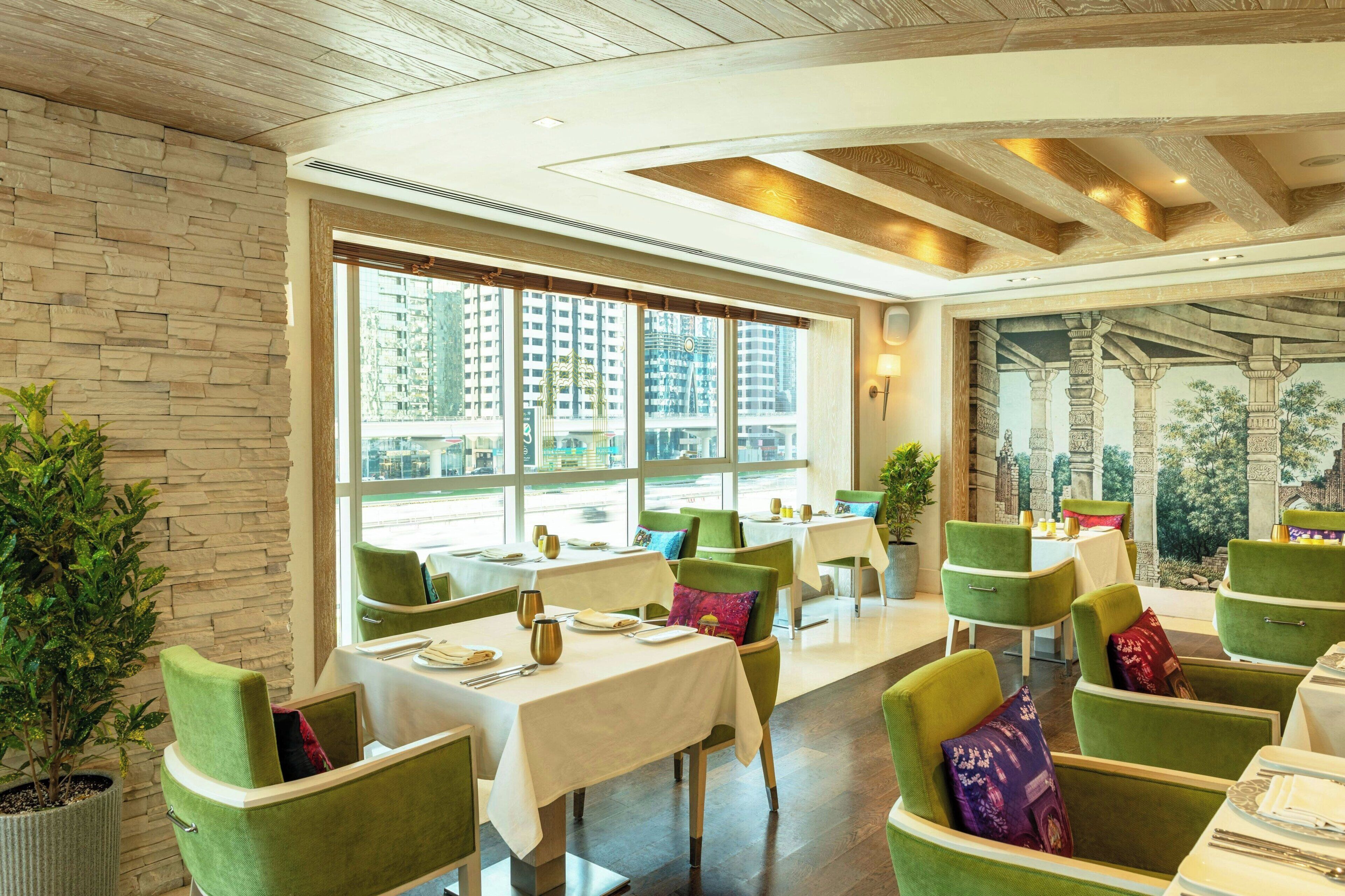 Un restaurant avec des chaises à revêtement vert, des nappes blanches et de grandes fenêtres donnant sur des immeubles.