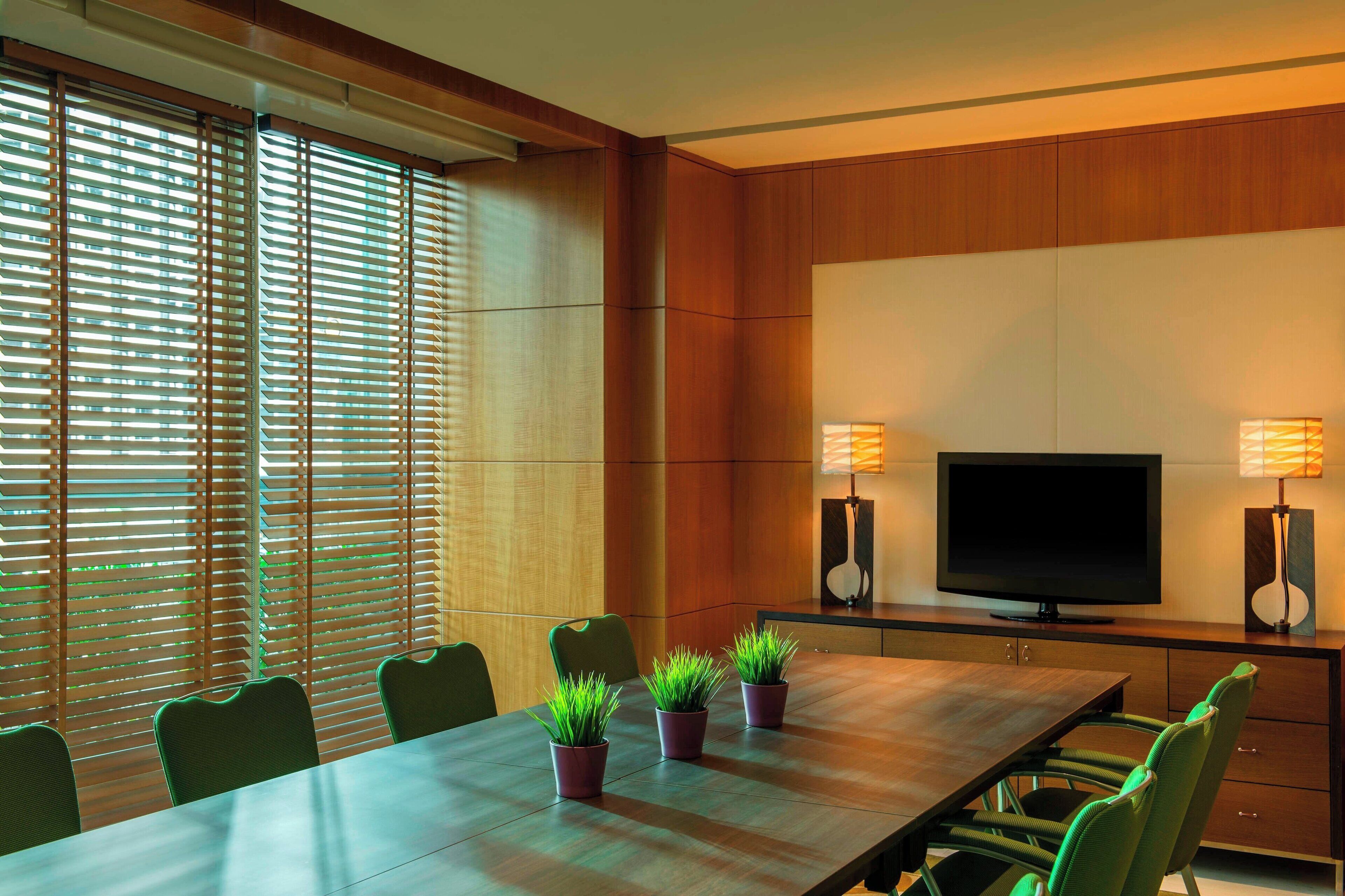 A modern conference room with a long table, green chairs, potted plants, a flat-screen TV, and two wall-mounted lamps.