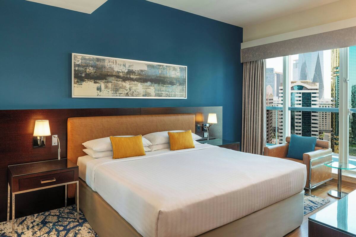 A hotel room with a large bed, two bedside tables with lamps, a chair, a glass table, and a city view through the window.