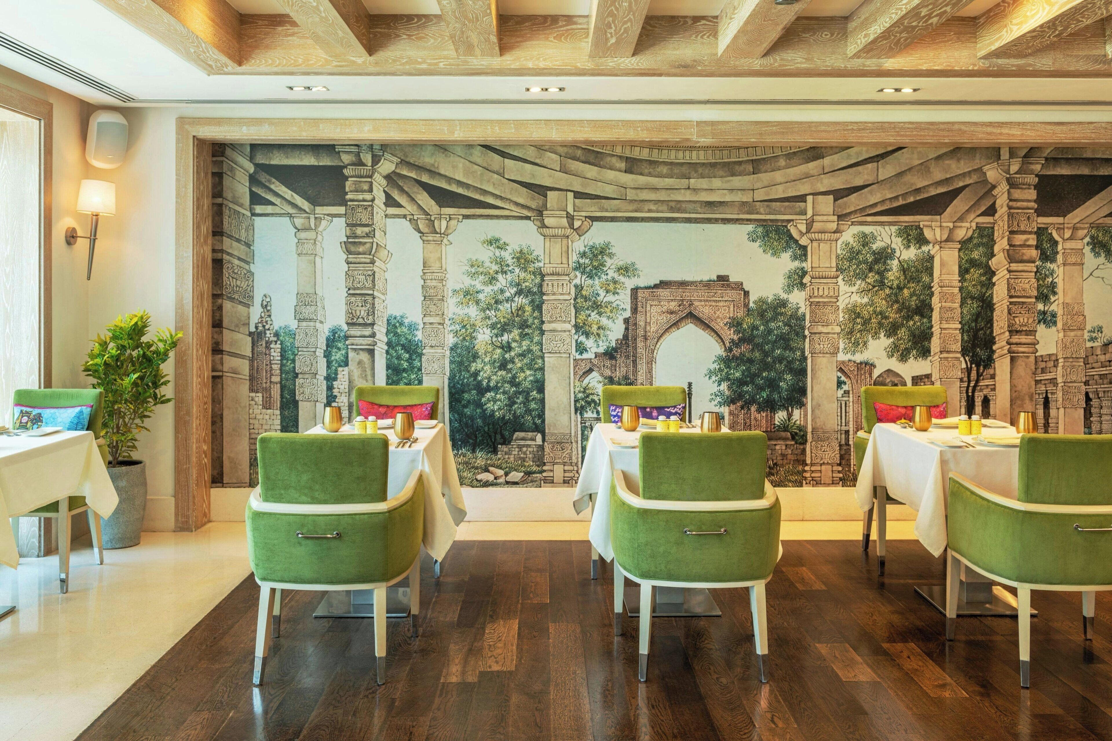 A restaurant with green chairs and white tables, a mural of an ancient structure, and a potted plant.