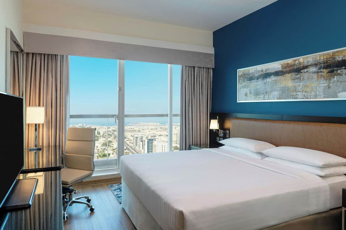 A hotel room with a large bed, a desk, a chair, and a view of the city through large windows.