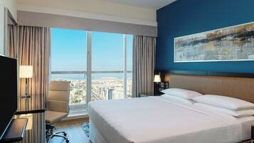 Habitación de hotel con una cama grande, un escritorio, una silla y vistas a la ciudad a través de amplios ventanales.