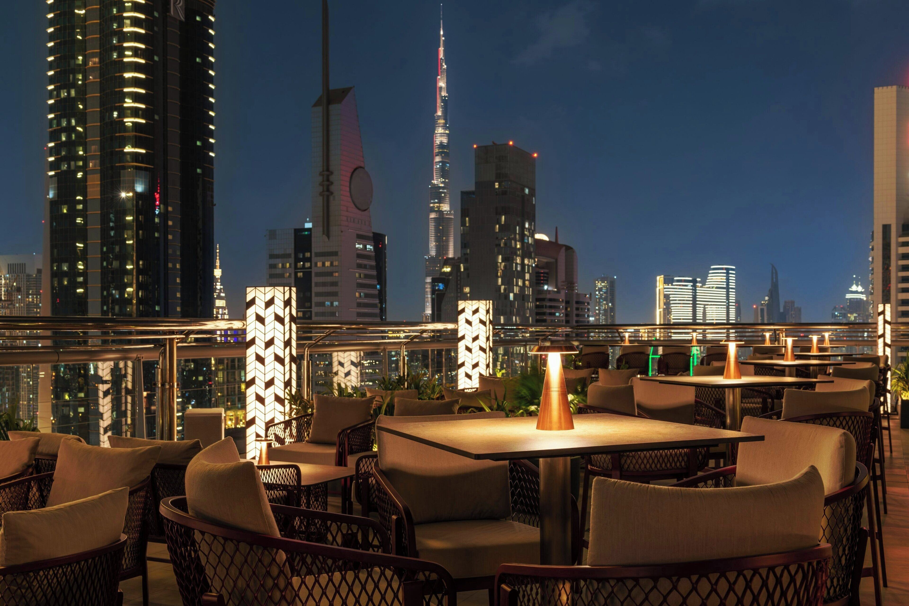 An outdoor dining area with tables and chairs, overlooking a city skyline at night.