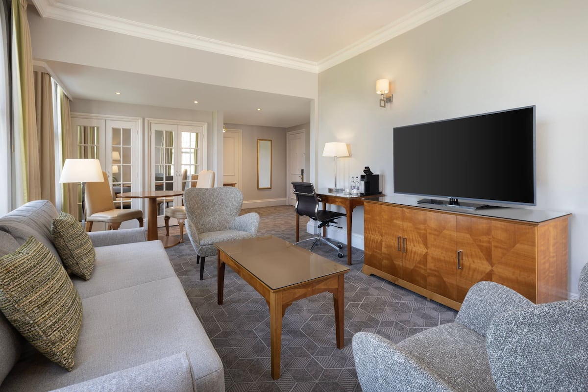 A hotel room with a sofa, armchairs, a wooden cabinet with a TV, and a desk with a lamp.